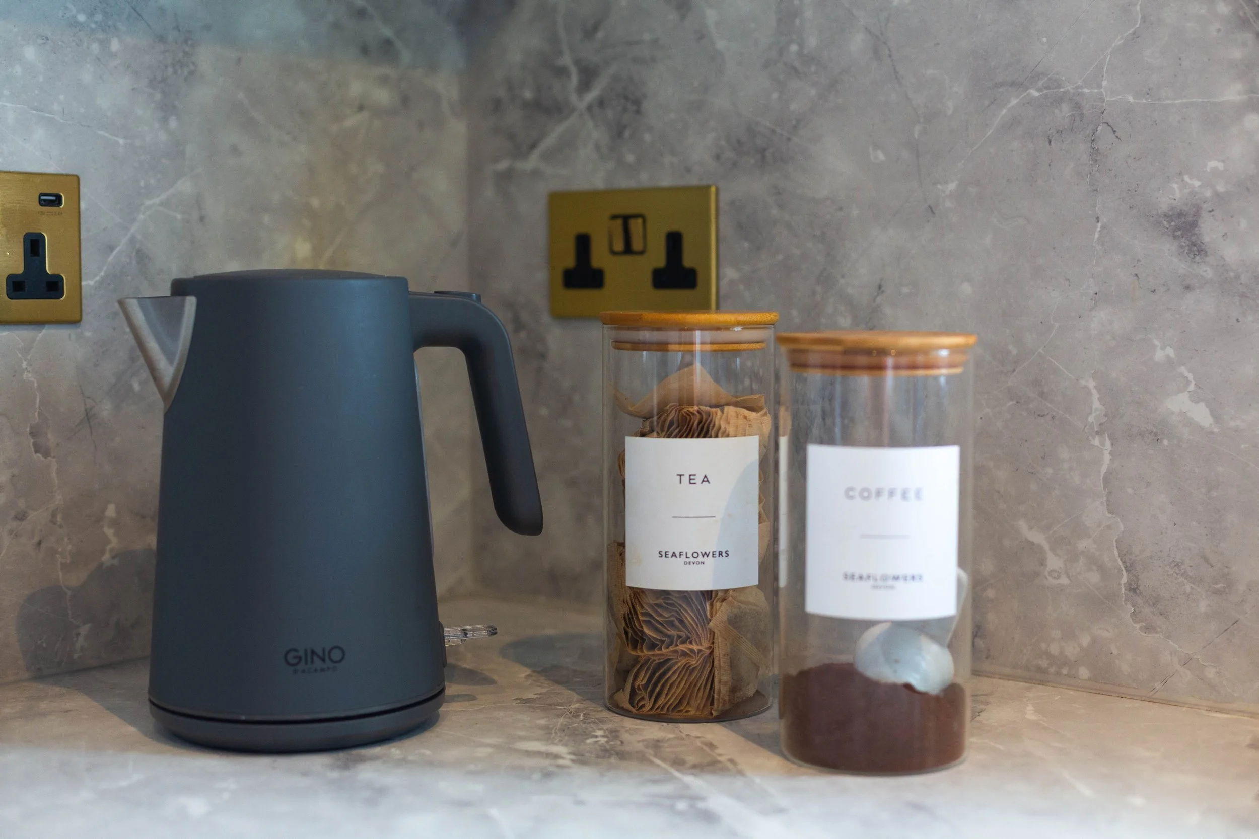 A matte gray electric kettle on a kitchen countertop, with two glass jars labeled 'Tea' and 'Coffee' beside it. The background features a marble wall with two yellow electrical outlets and a black electrical plug symbol.