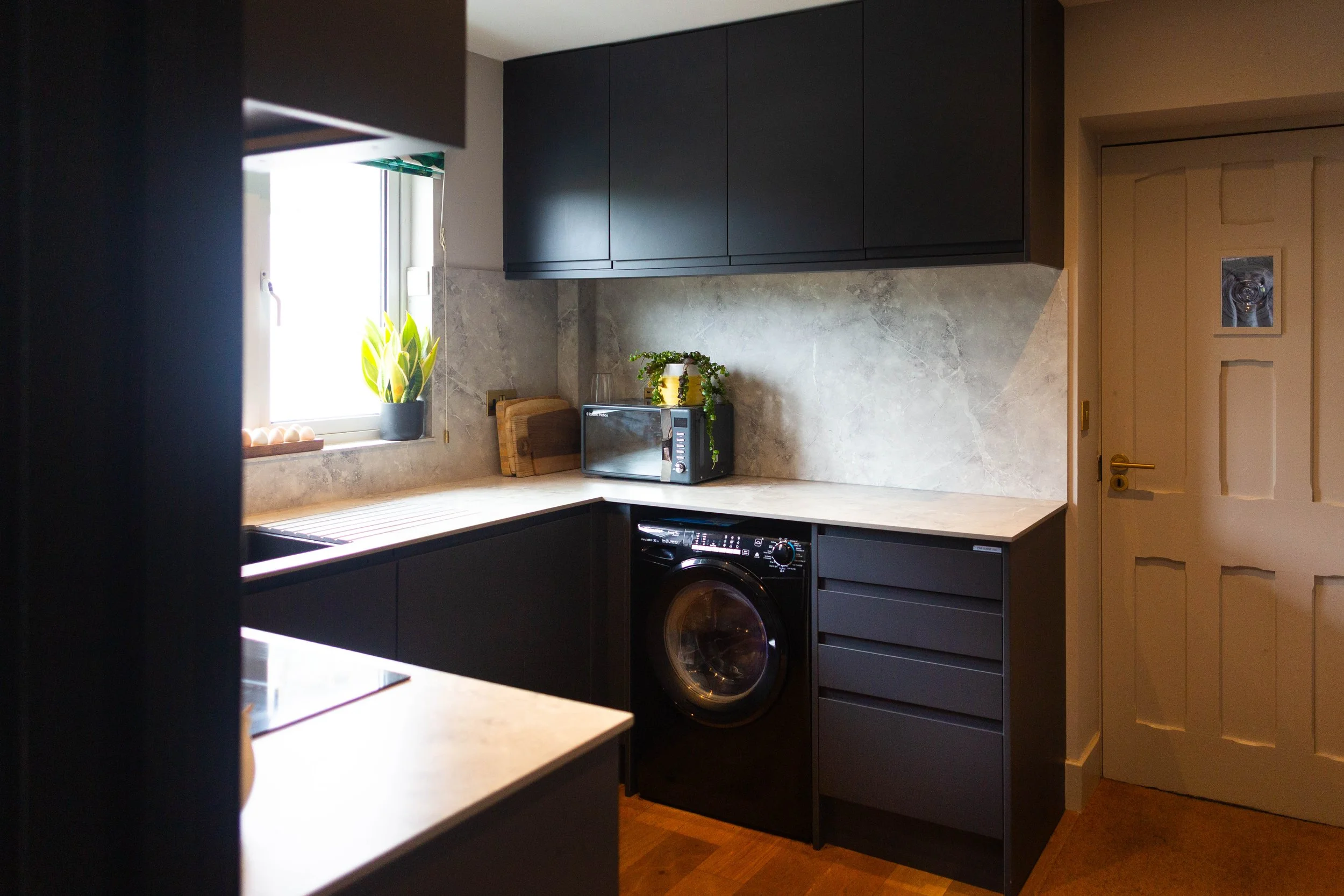 A kitchen with dark cabinets, a window with a potted plant, a microwave, and a washing machine.