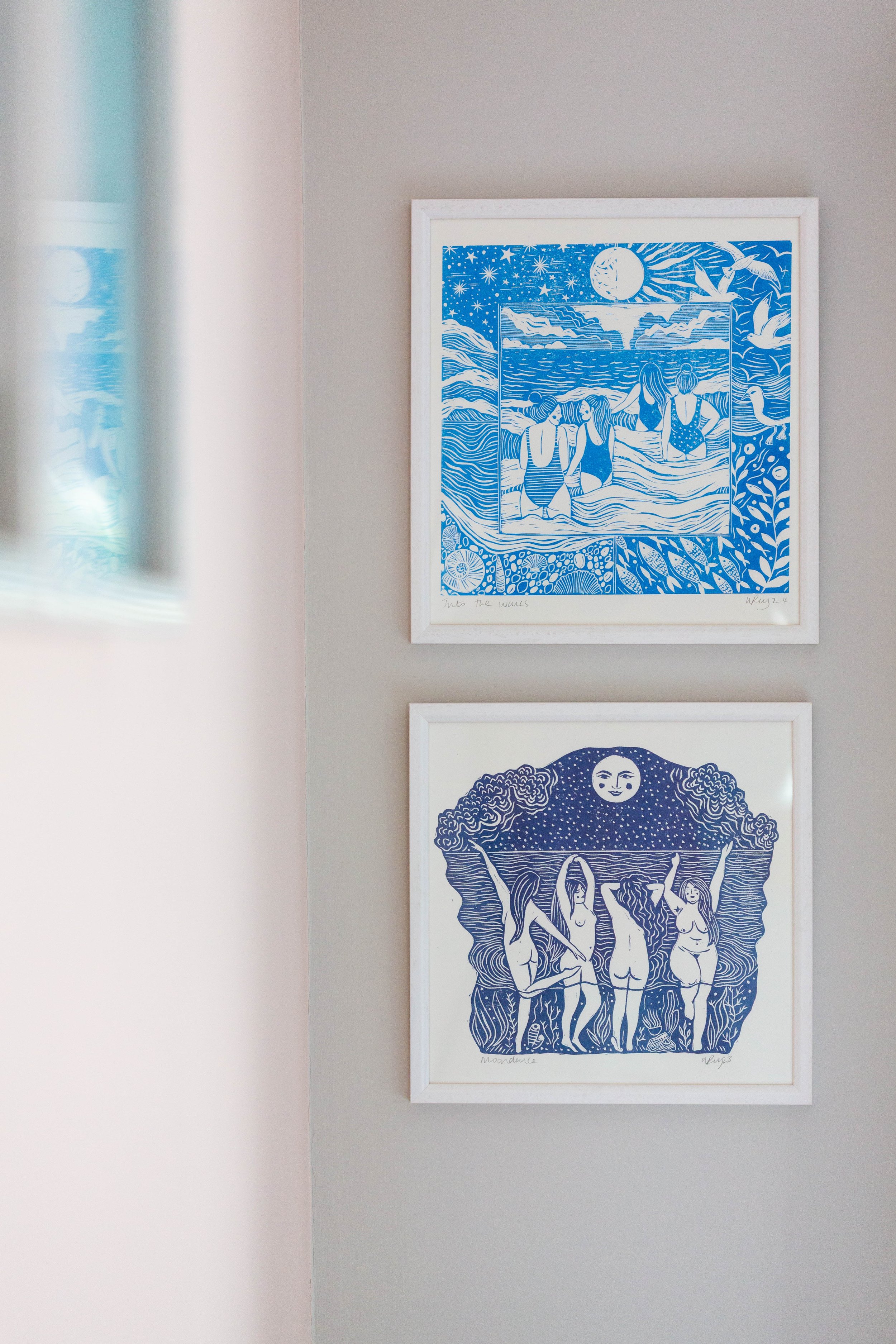 Two framed art prints of blue and white illustrations. The top print depicts four women in swimsuits wading in the water at sunset or sunrise with seagulls flying overhead. The bottom print features five women with arms raised, surrounded by an intri