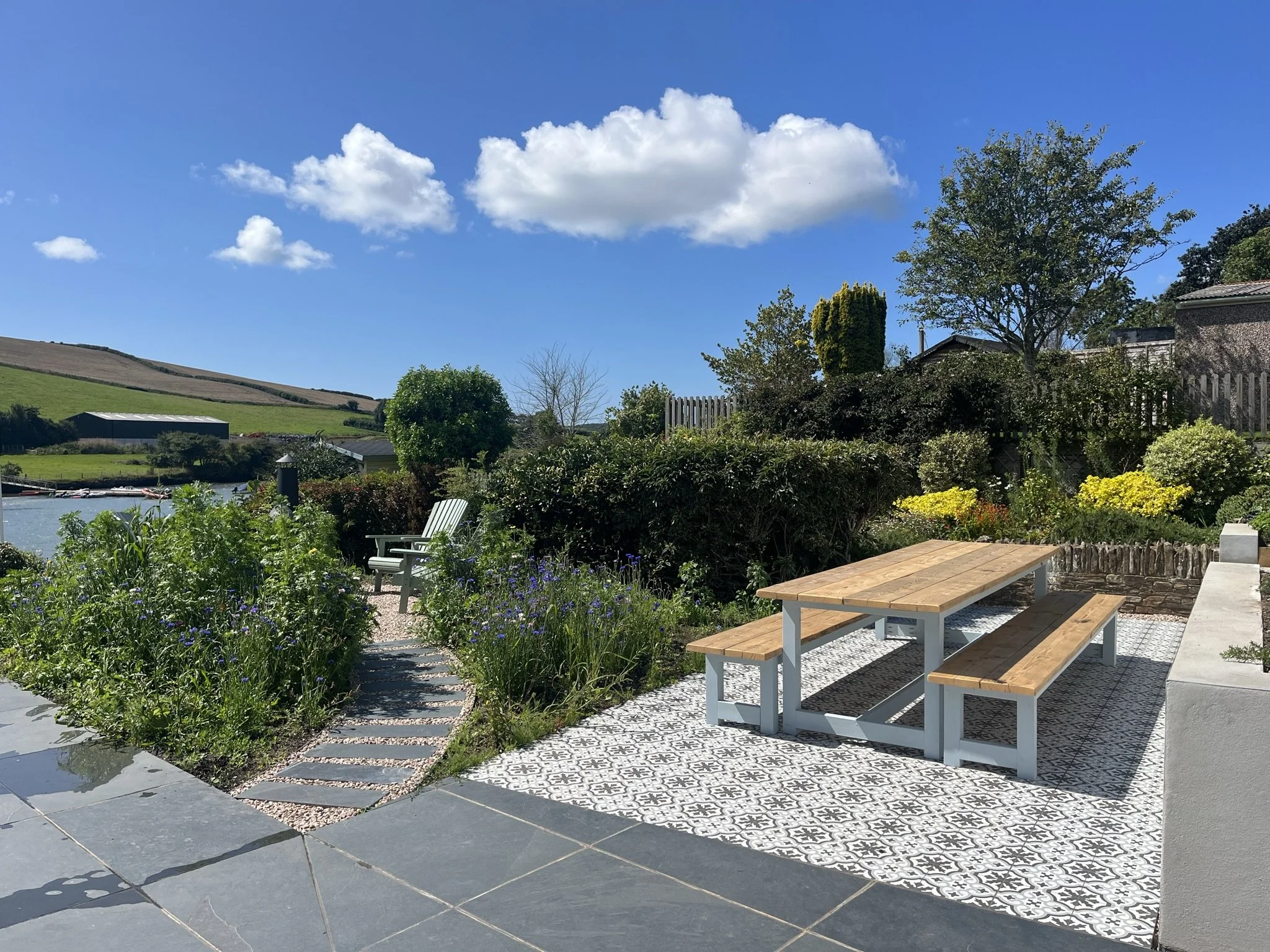 A backyard with a stone patio, wooden benches, plants, trees, and a view of rolling hills under a blue sky with white clouds.