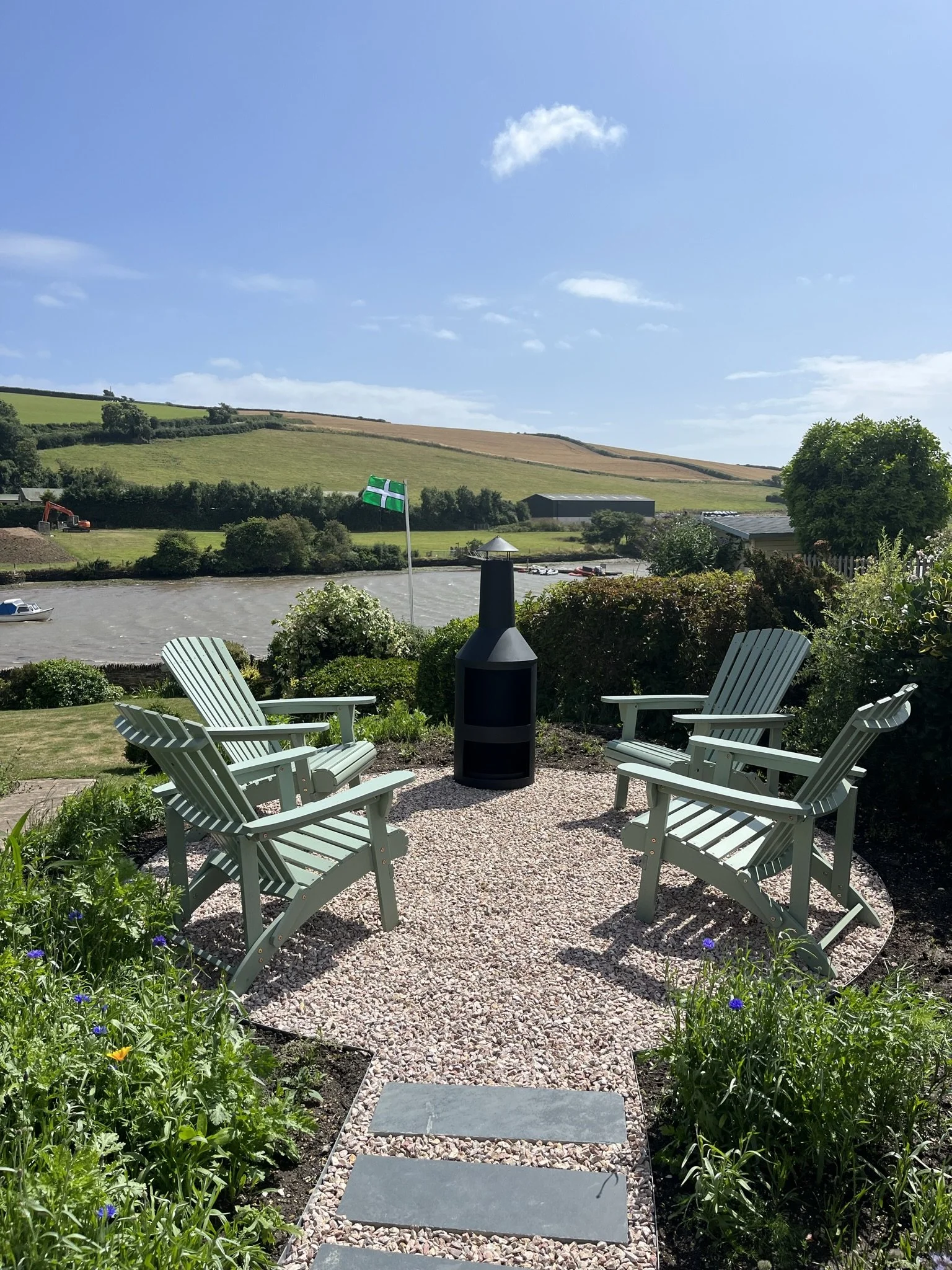 Six white Adirondack chairs arranged in a circle on a gravel patio surrounded by greenery, with a small fire pit in the center. In the background, a flagpole with a green and white flag, trees, a parking lot, and rolling fields under a partly cloudy 