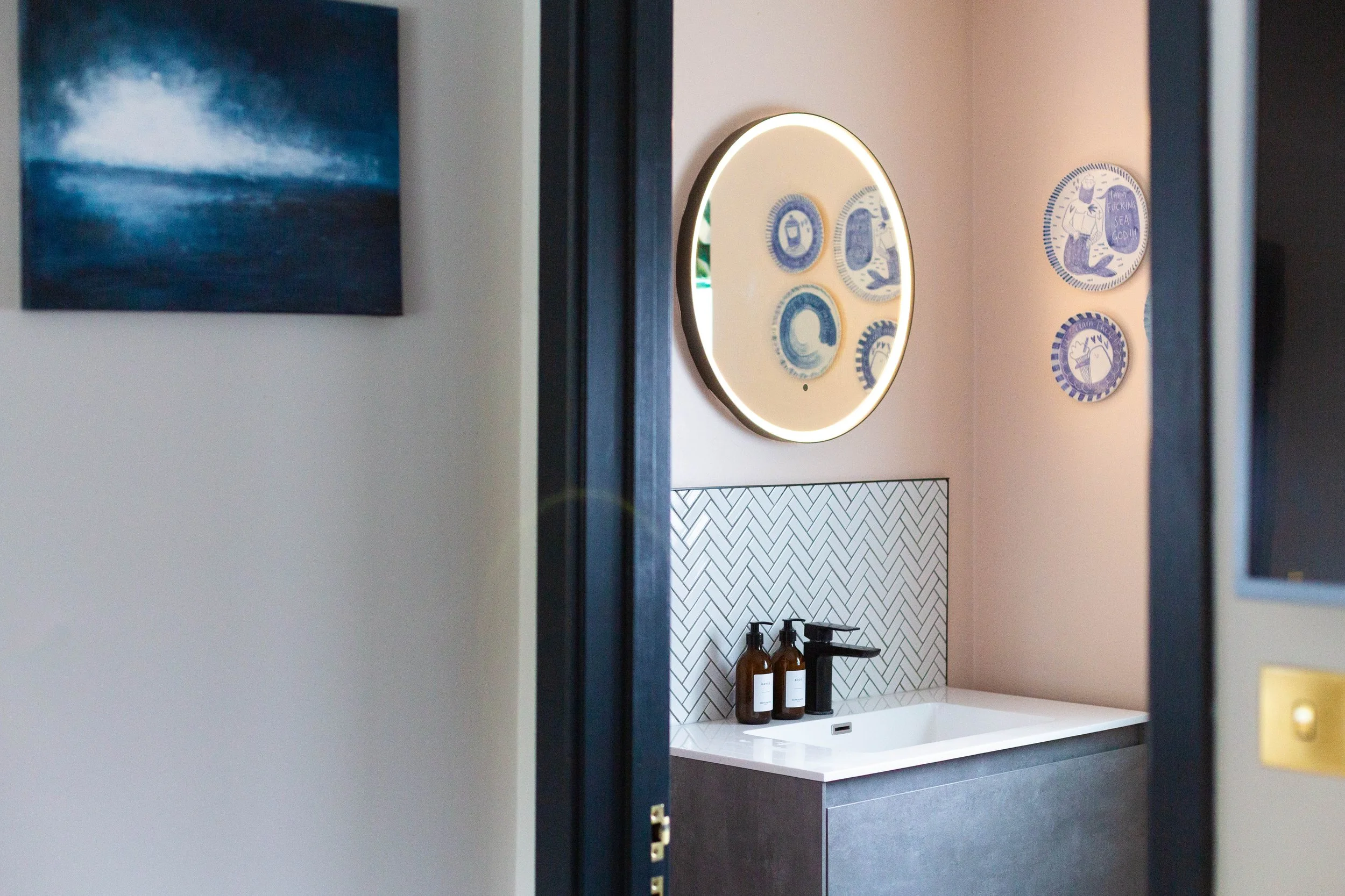 A bathroom vanity with a white countertop, a black faucet, and two amber bottles. Backsplash with a herringbone tile pattern, large round wall mirror with a lighted frame, and decorative art pieces on the wall.