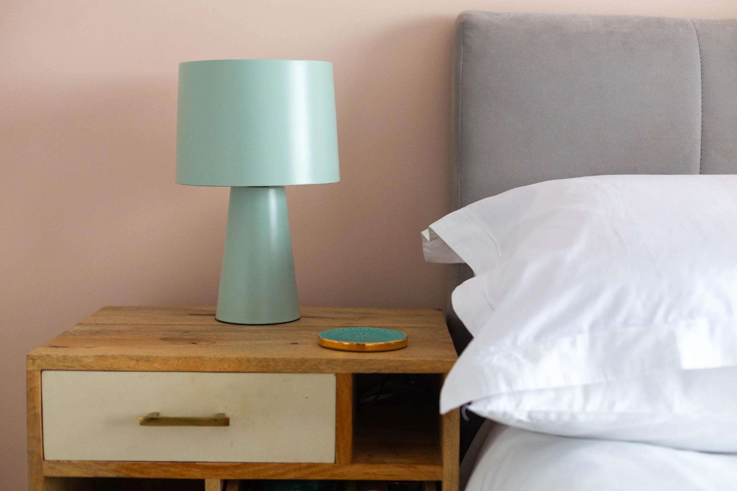 A wooden nightstand with a white drawer and handle, a mint green lamp, and a decorative plate with green and orange colors next to a bed with white sheets, against a light pink wall.