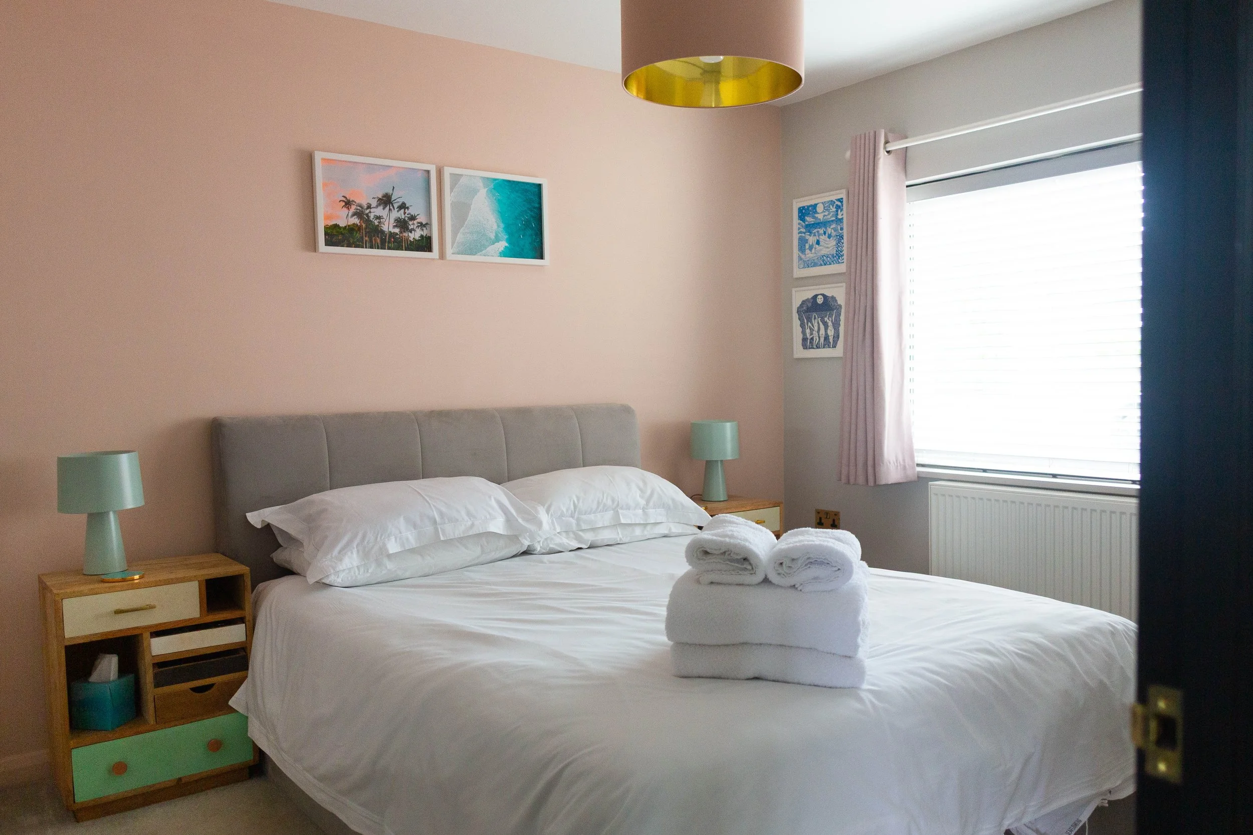 A neatly made bed with white linens and towel roll preparation, flanked by wooden nightstands with turquoise and pastel drawer fronts, in a bedroom with pastel pink and gray walls, featuring framed artwork including beach scenes and abstract art, a w