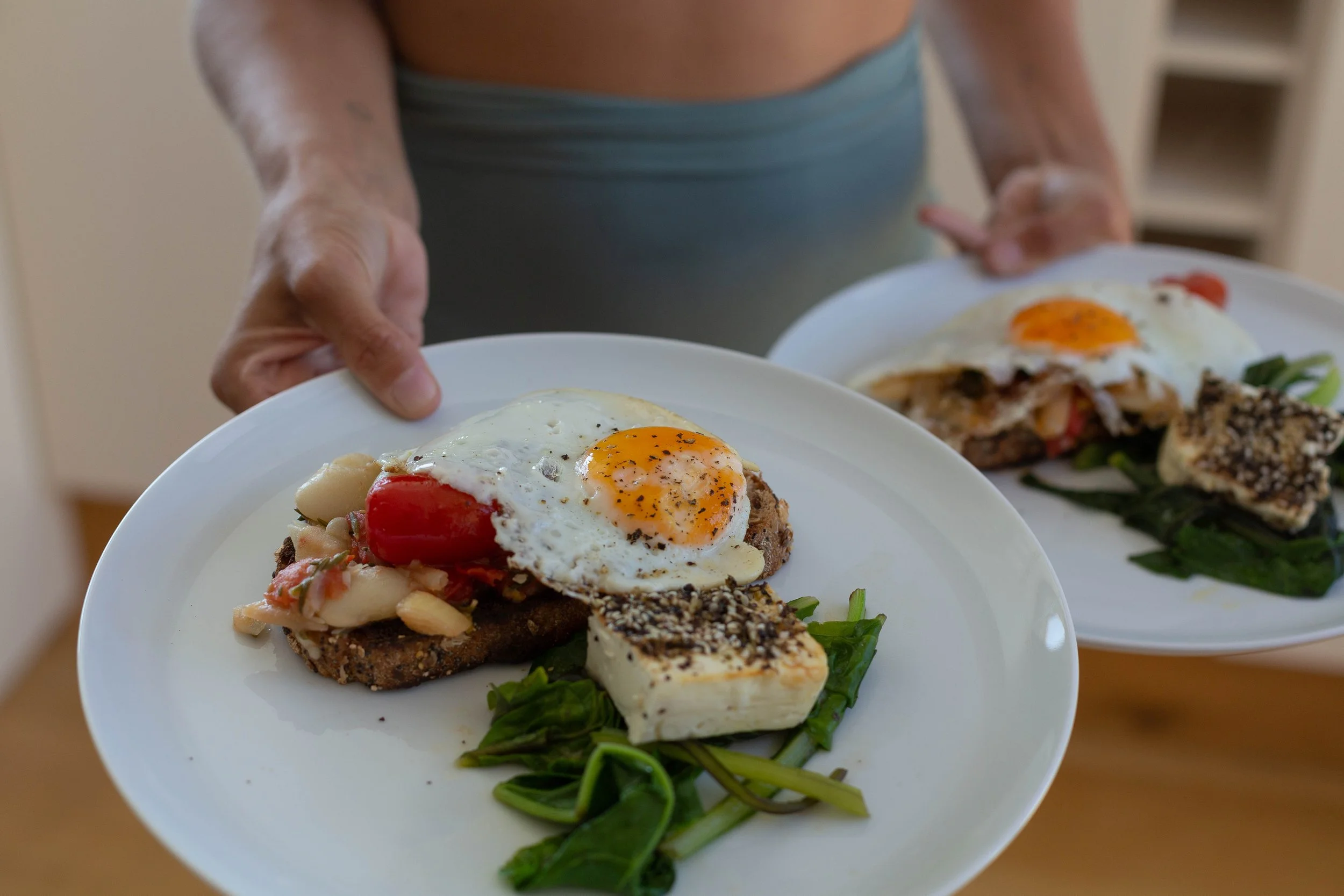 Person holding white plates with breakfast consisting of fried eggs, grilled tomatoes, greens, feta cheese, and bread slices.