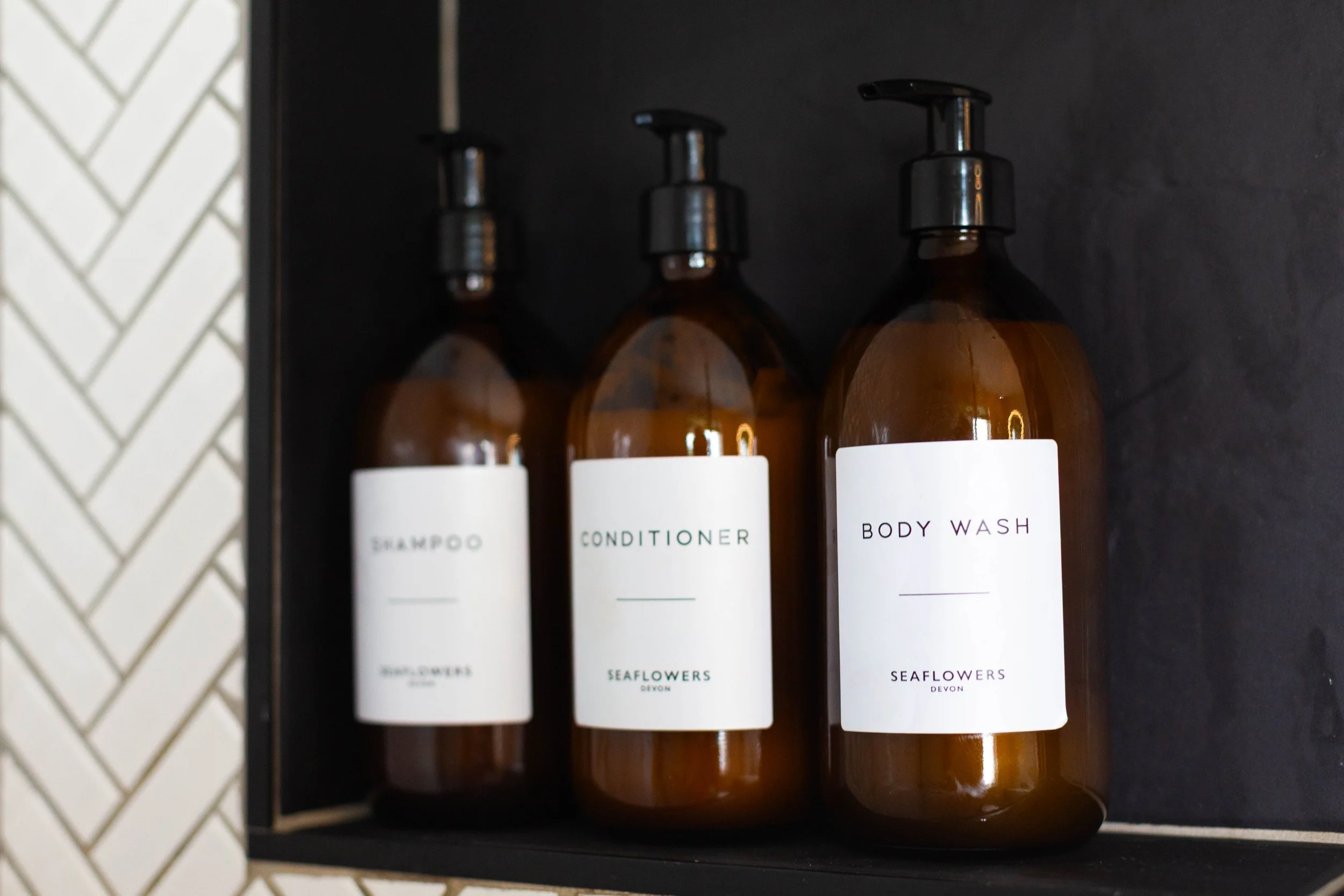 Three amber-colored bottles labeled shampoo, conditioner, and body wash are placed inside a black shelf with a geometric tiled wall in the background.