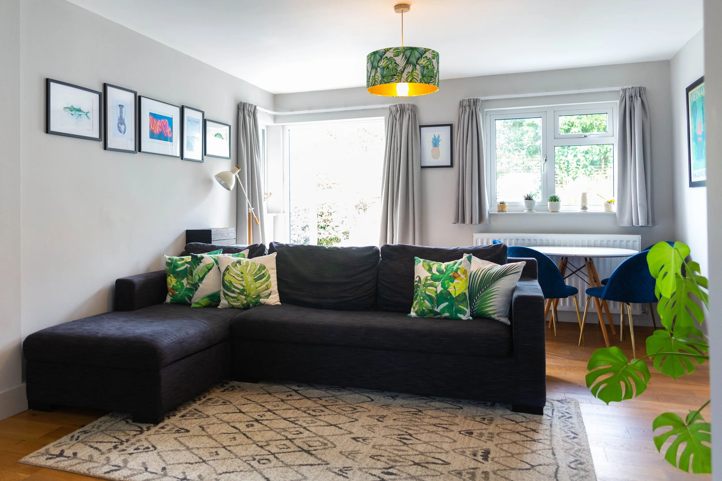 Living room with black sofa decorated with patterned pillows and a blanket, wooden floor, white walls with framed pictures, a dining table with blue chairs, large windows with curtains, a sliding glass door, green plants, and a ceiling light with tro