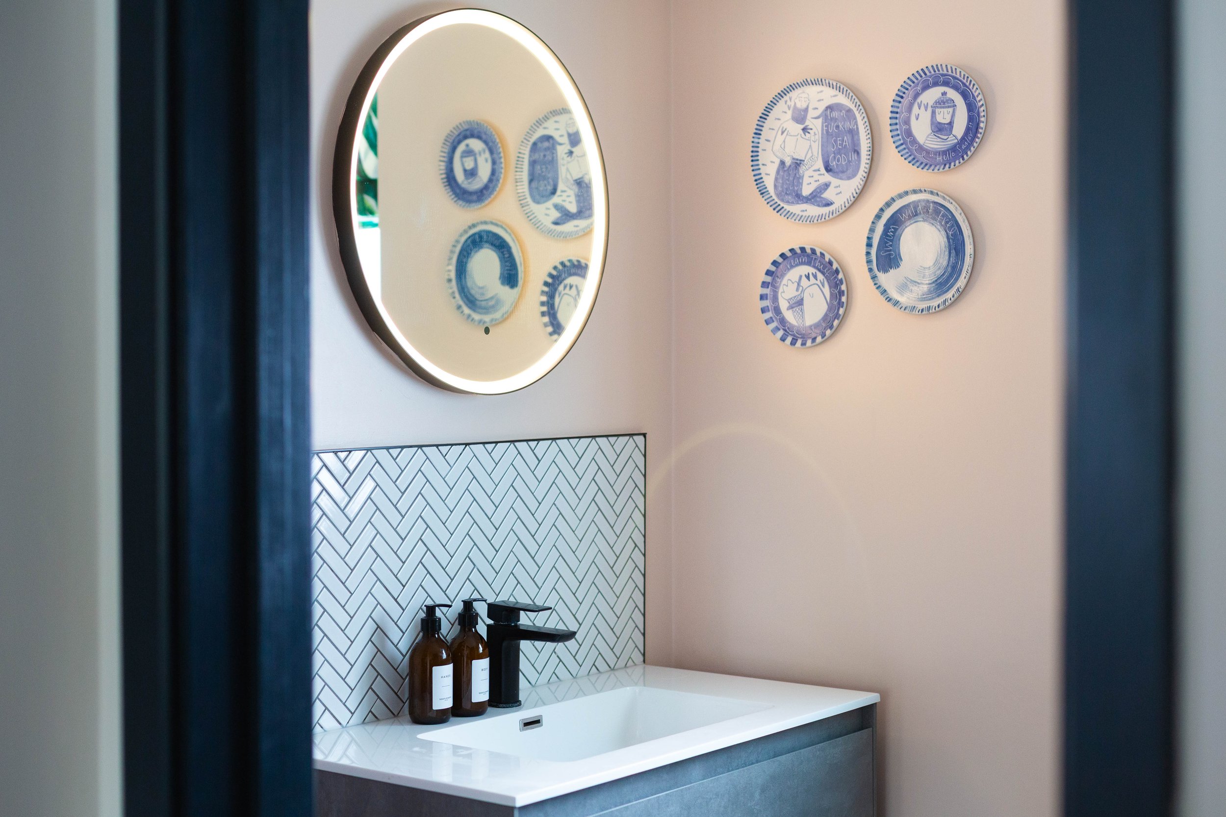 A modern bathroom vanity with a rectangular sink, a black faucet, and brown bottles on the countertop. Behind, there is a wall with white herringbone tiles and a round mirror with LED backlighting. To the right, there are six round wall decorations w