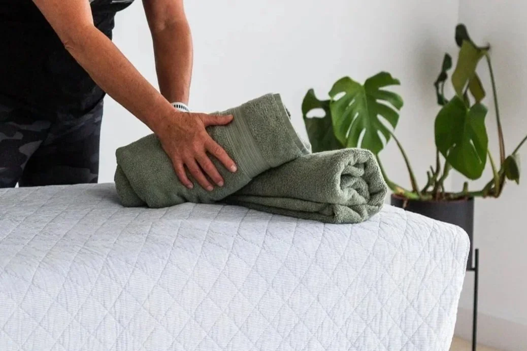 Person folding a green towel on a bed with a white quilted bedspread, next to a plant with large green leaves.