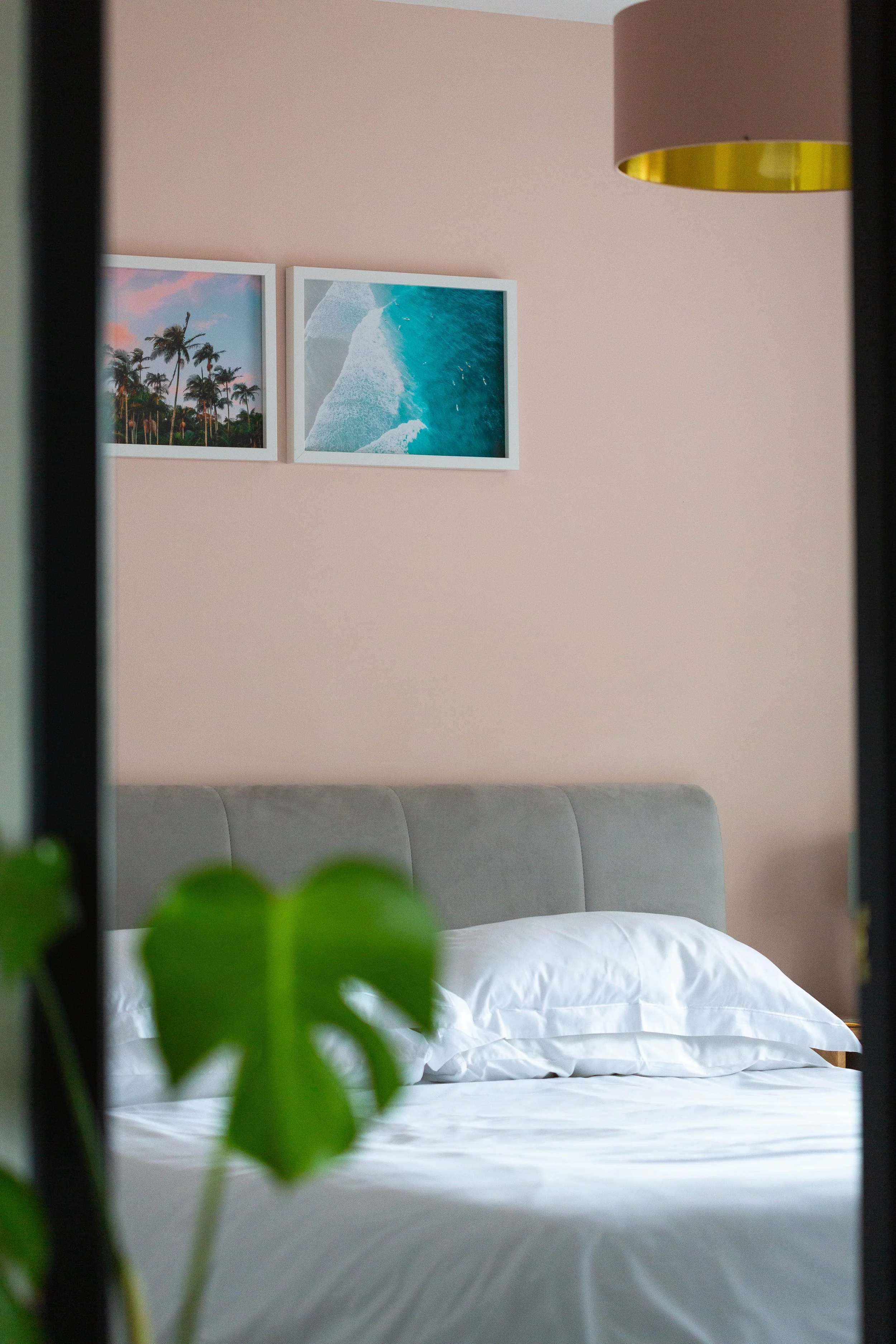A neatly made bed with white bedding and pillows, positioned against a pale pink wall decorated with two framed photographs of coastal scenery. A small plant with green leaves is in the foreground, partially obscuring the view.