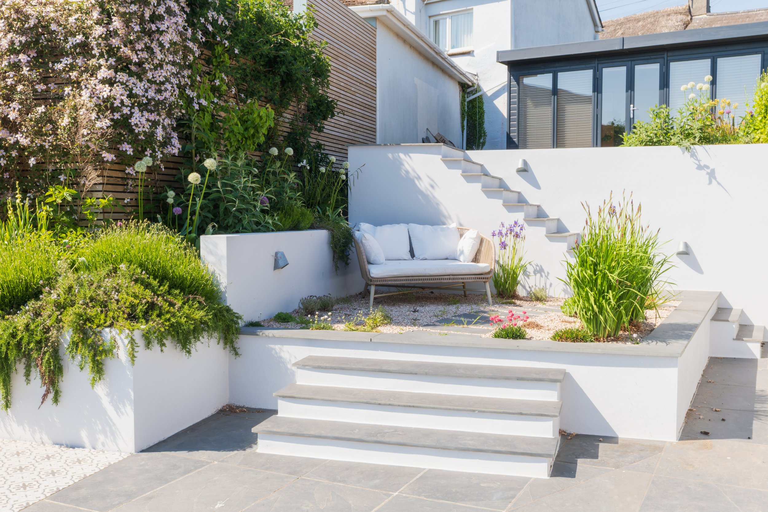 A modern outdoor patio with white stairs leading up to a raised garden area. The garden is lush with various plants and flowers, including a large flowering shrub on the left and potted plants on the right. A white bench with cushions is positioned a