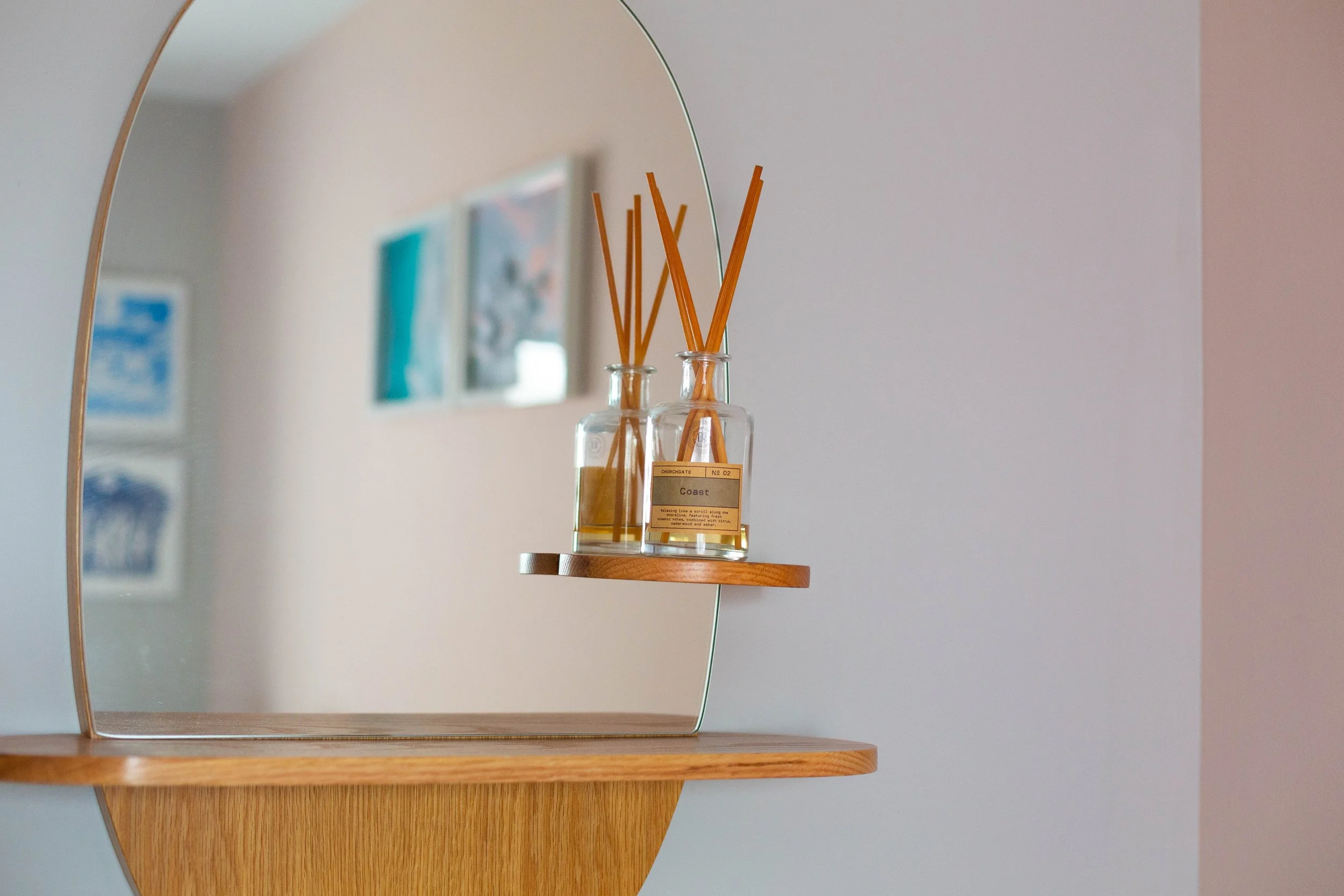 A mirror reflecting a wooden shelf with a glass bottle of reed diffuser sticks labeled 'Coast' and some artwork on the wall in the background.