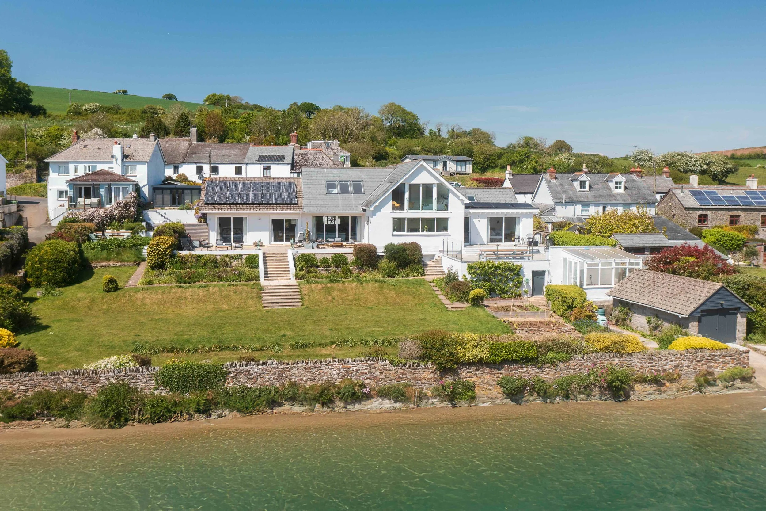 A large white house on a hill next to a body of water, with a well-kept garden and steps leading down to the water.