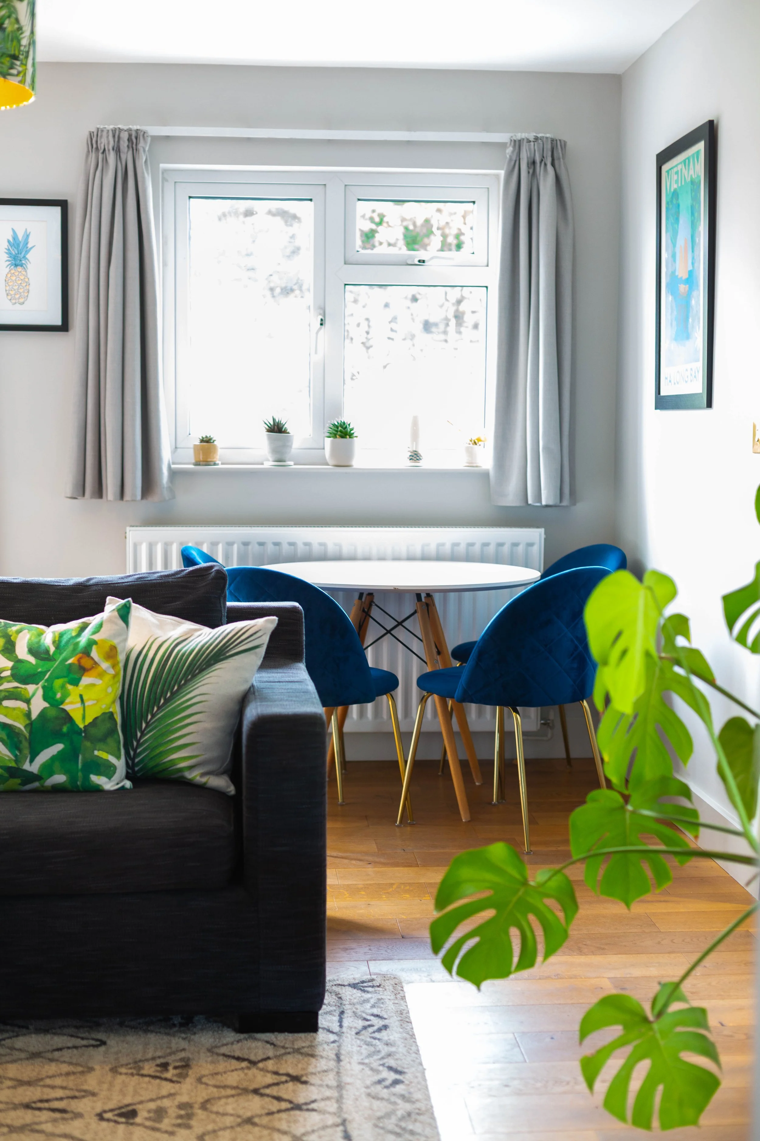 Living room with black sofa decorated with patterned pillows and a blanket, wooden floor, white walls with framed pictures, a dining table with blue chairs, large windows with curtains, a sliding glass door, green plants, and a ceiling light with tro