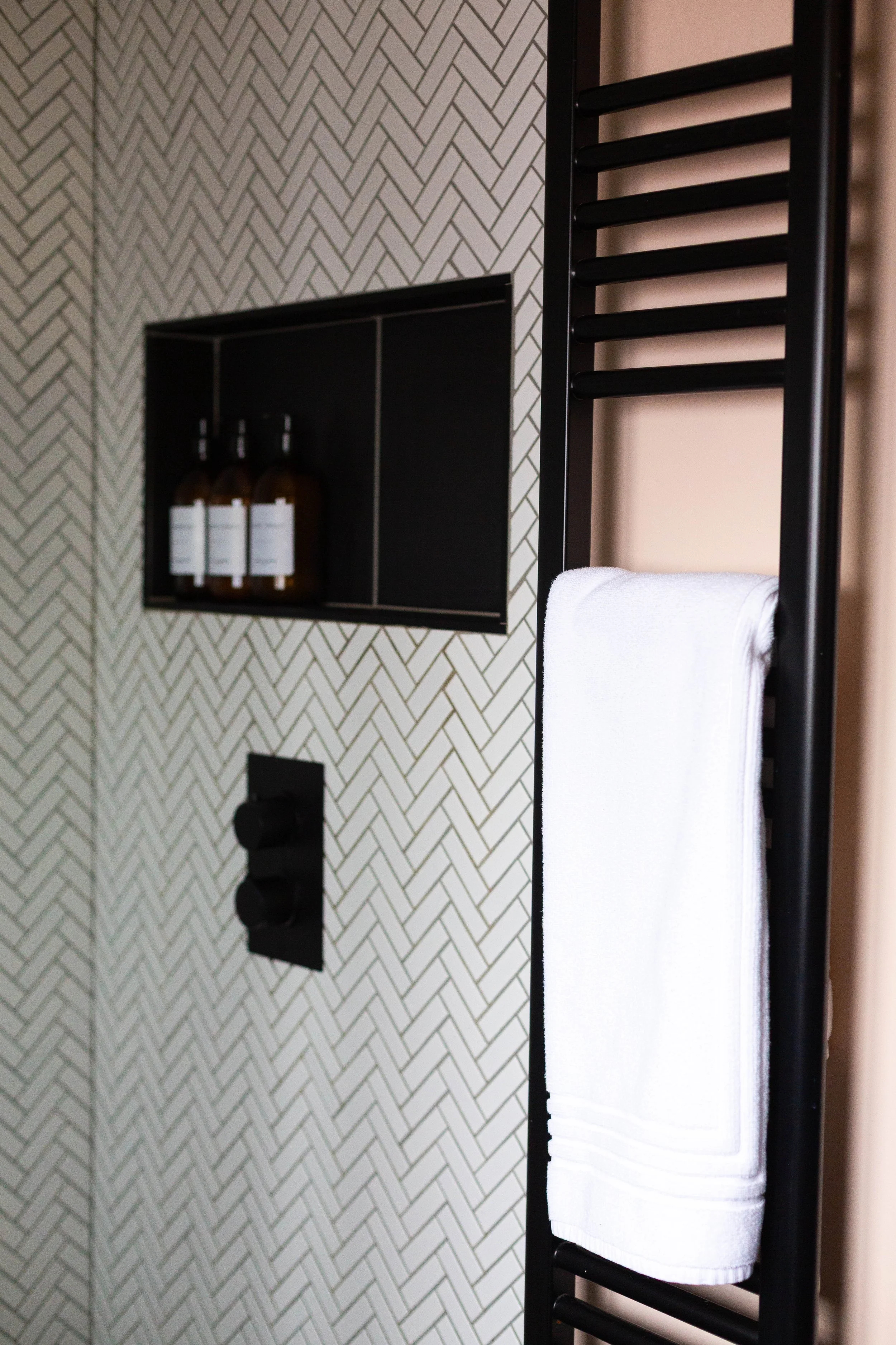 A black towel rack with a white folded towel hanging on it, positioned next to a wall with a black and white herringbone pattern tile design, and a black wall-mounted bottle holder with three bottles.