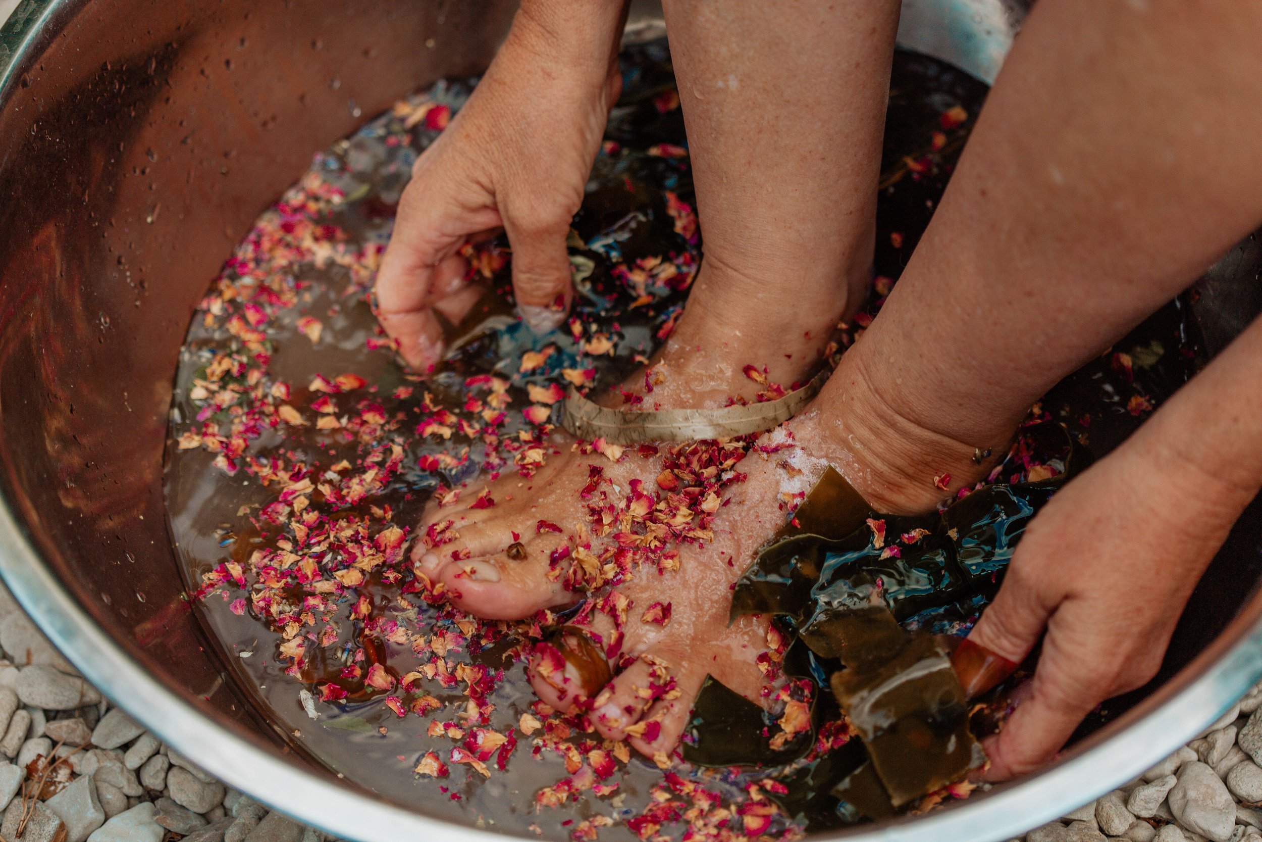 Close-up of a person's feet soaking in a water basin with flower petals, with a hand touching one foot, and two metal circular foot soak rings, on a gravel ground.