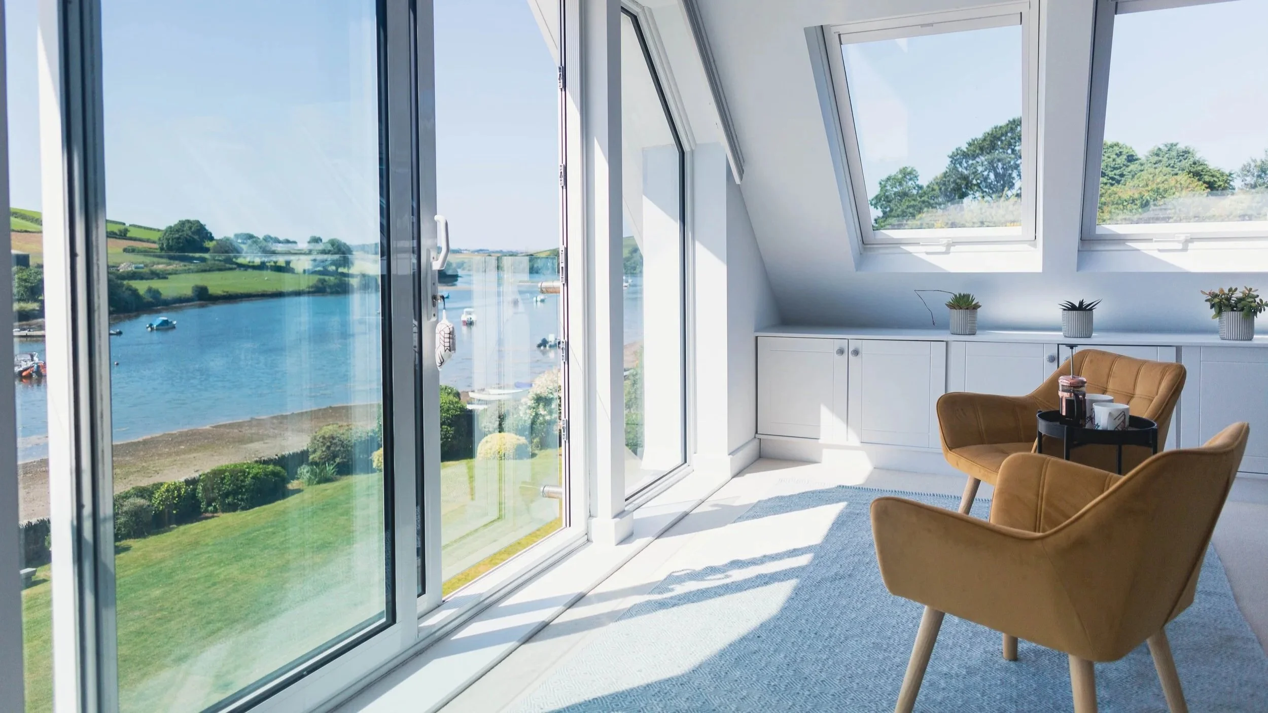Bright room with large windows overlooking water and green hills, furnished with two yellow armchairs, a small black side table, and a bed partially visible on the left.