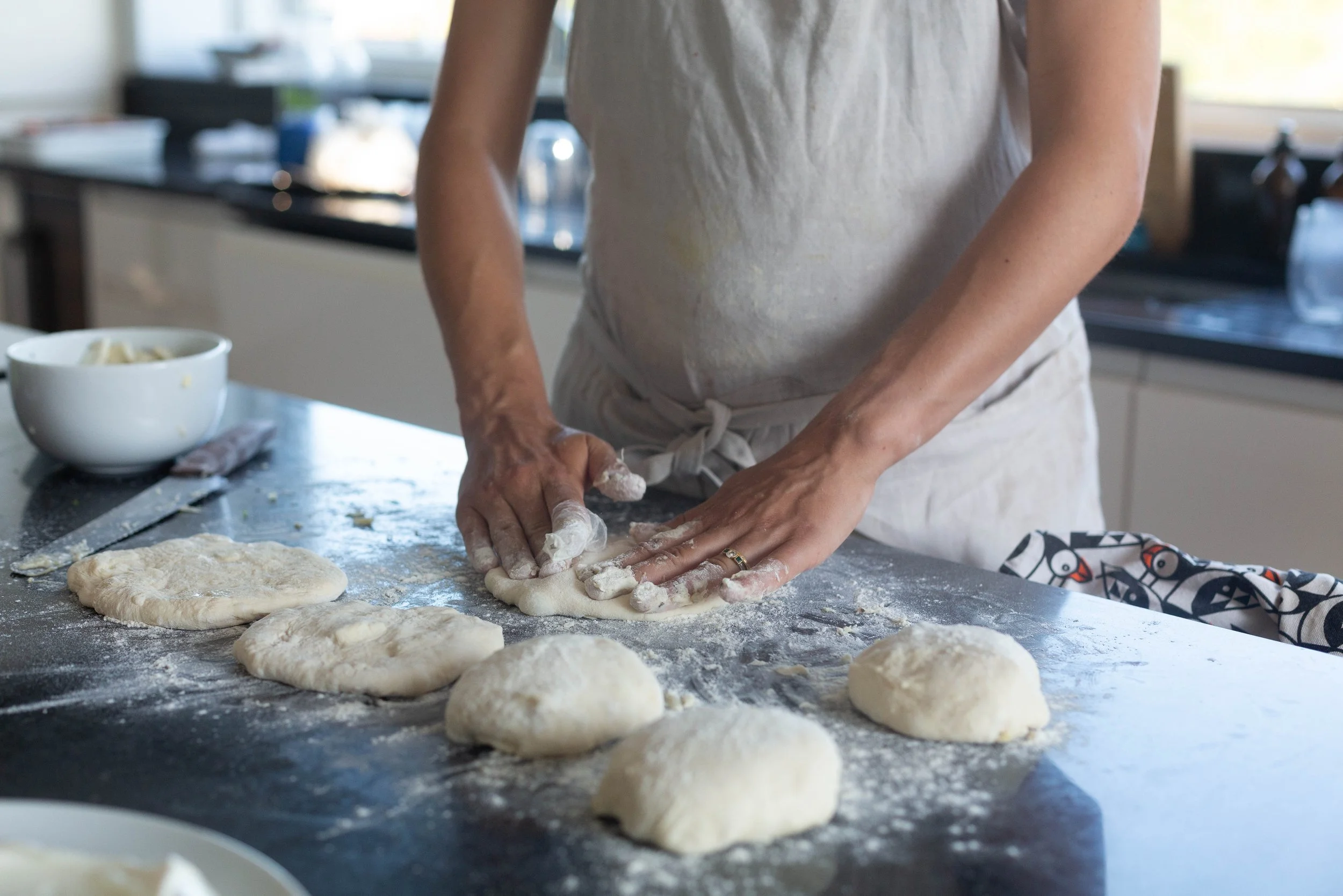 Person in a white apron kneading dough on a floured kitchen counter with several pieces of dough shaped into rounds and a bowl of ingredients in the background.