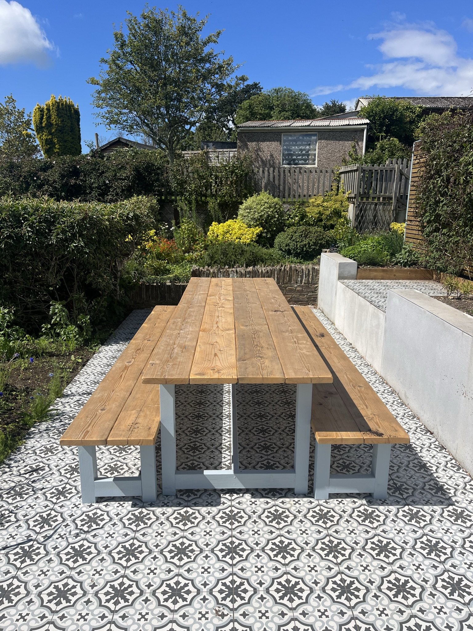 A wooden picnic table and benches on a decorative black and white tiled patio in a backyard garden with bushes, trees, a wooden fence, and a small building with a window.