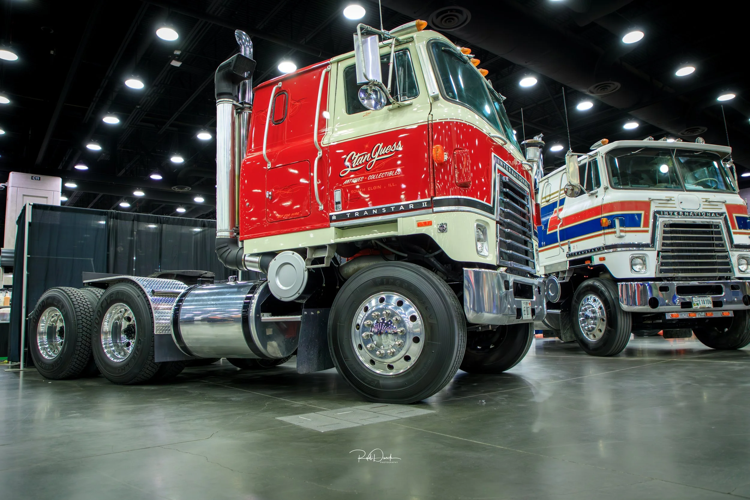 Classic semi-trucks on display indoors at an automotive show, featuring a red and white cab-over model.