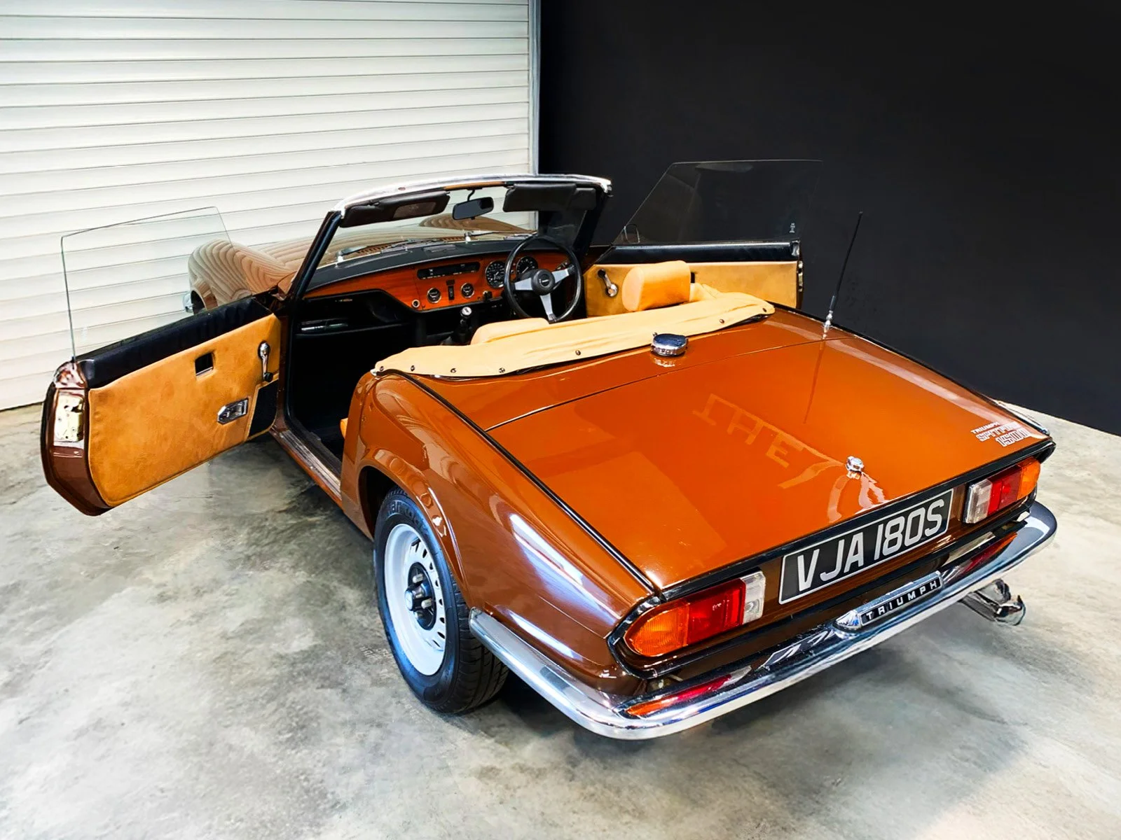 Vintage brown Triumph convertible car with open doors, tan interior, and a spare tire cover, parked indoors with black and white walls.