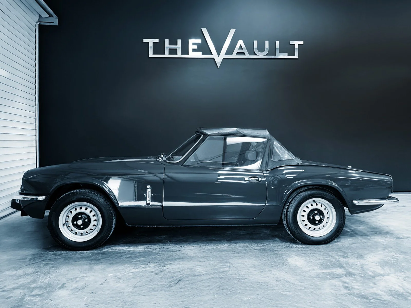 Black vintage convertible sports car parked indoors in front of a dark wall with a sign that says 'The Vault'.
