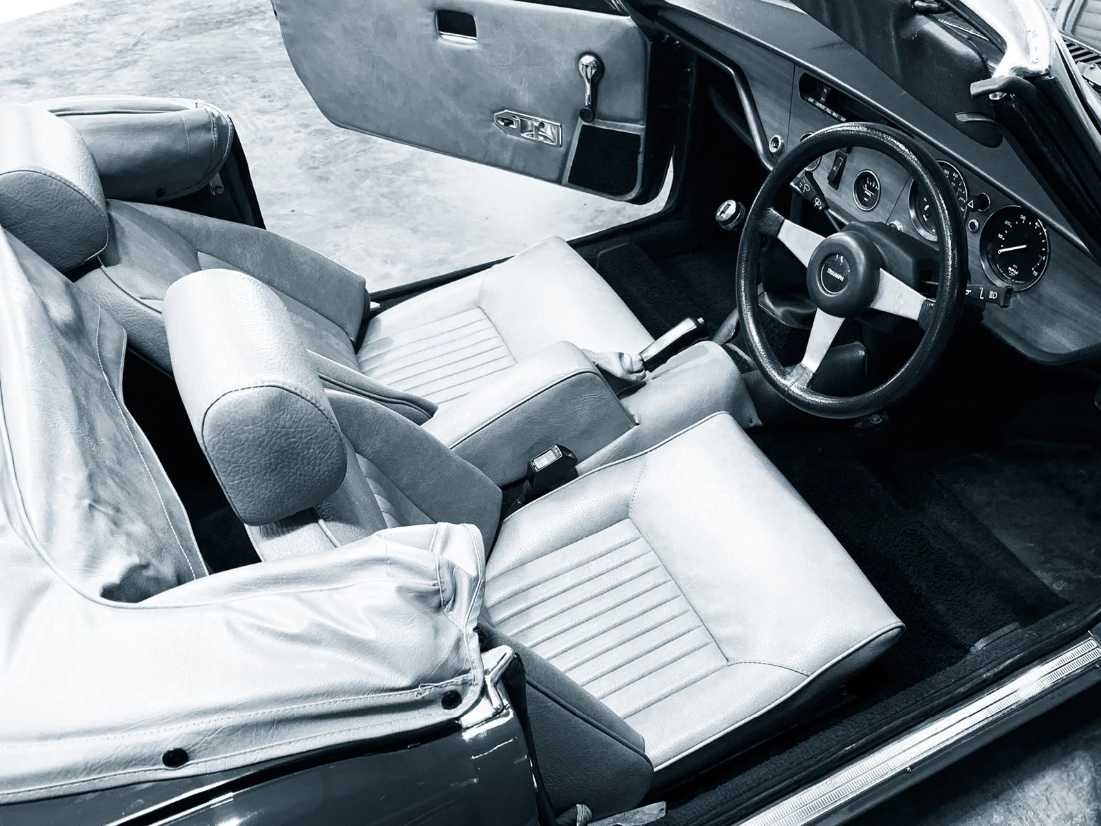 The interior of a vintage car showing the driver's seat, steering wheel, dashboard with gauges, and part of the passenger seat.
