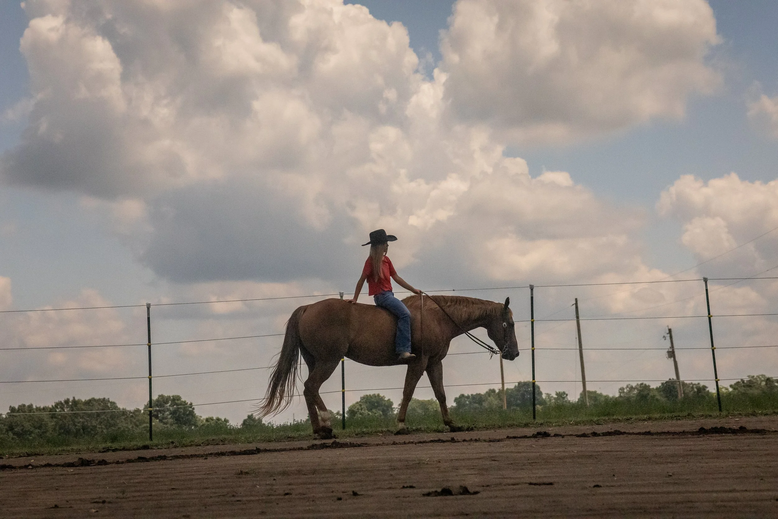 A person wearing a red shirt, jeans, and a black hat riding a horse along a fenced pasture with trees and cloudy sky in the background.