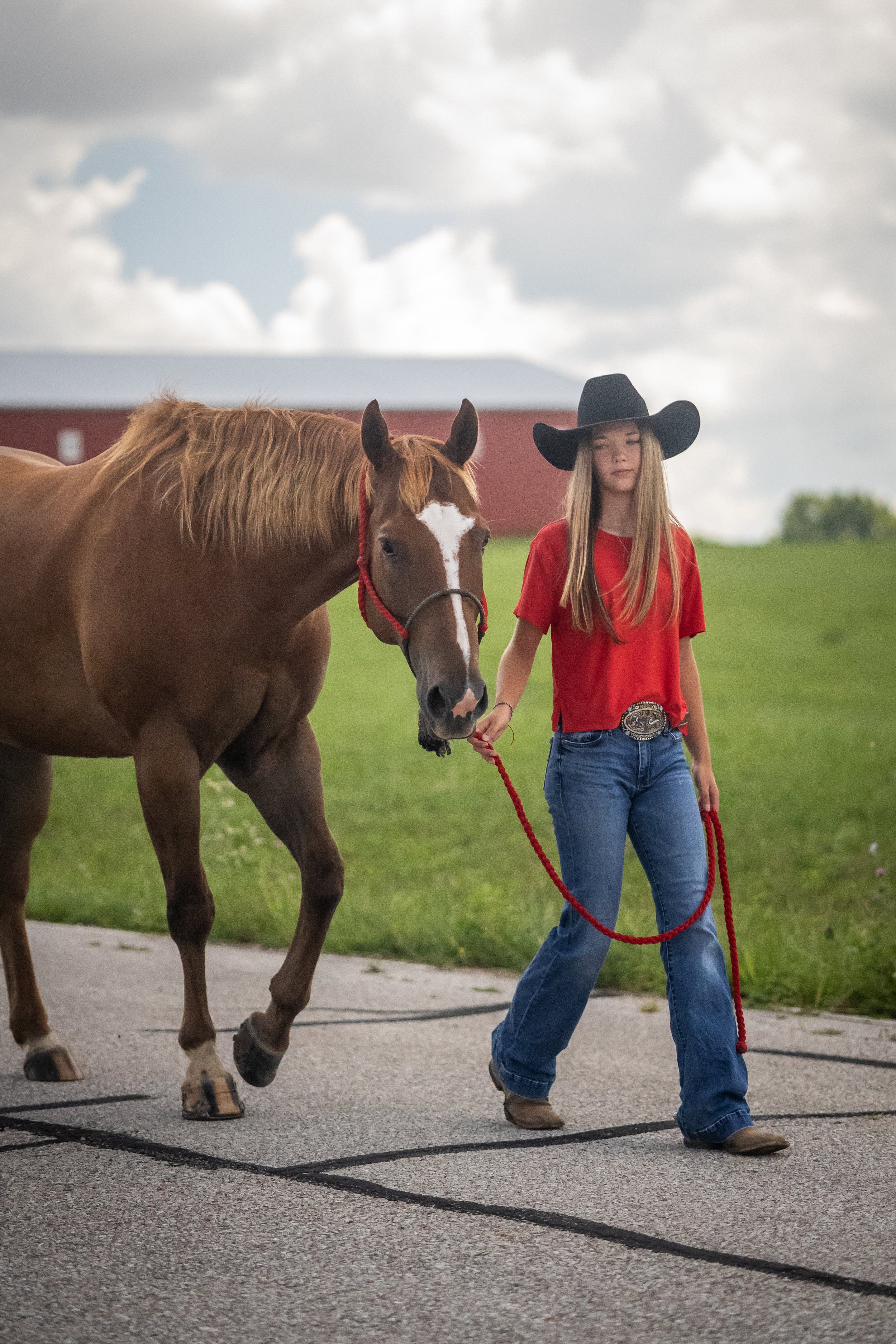 A young woman wearing a large black cowboy hat, red shirt, and blue jeans walking a brown horse with a white star on its face on a paved path in a rural area with a green field and a red barn in the background.