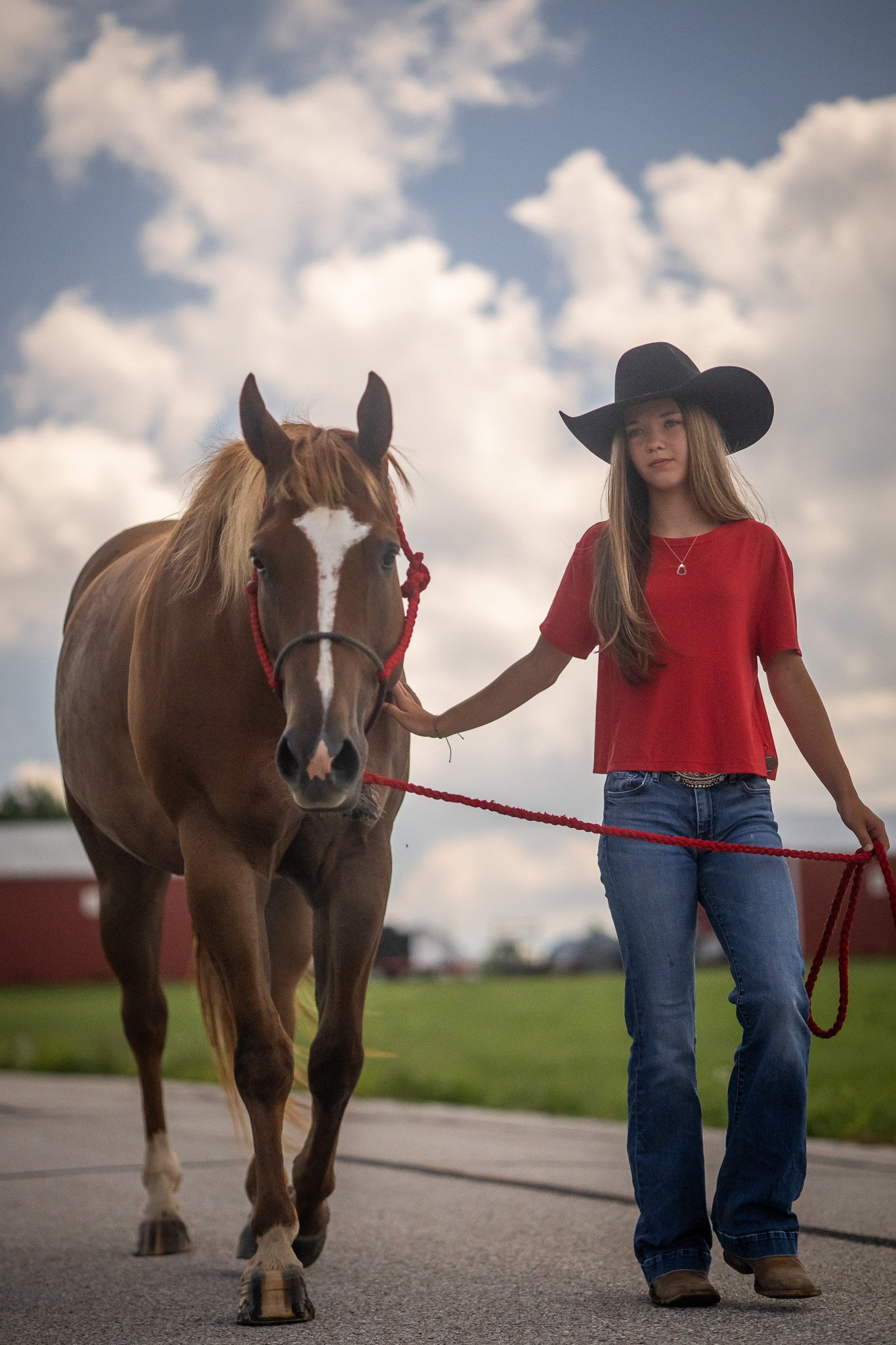 A young woman wearing a black wide-brimmed hat, a red short-sleeved shirt, and jeans walking a brown horse on a leash during daytime, with a cloudy sky in the background.