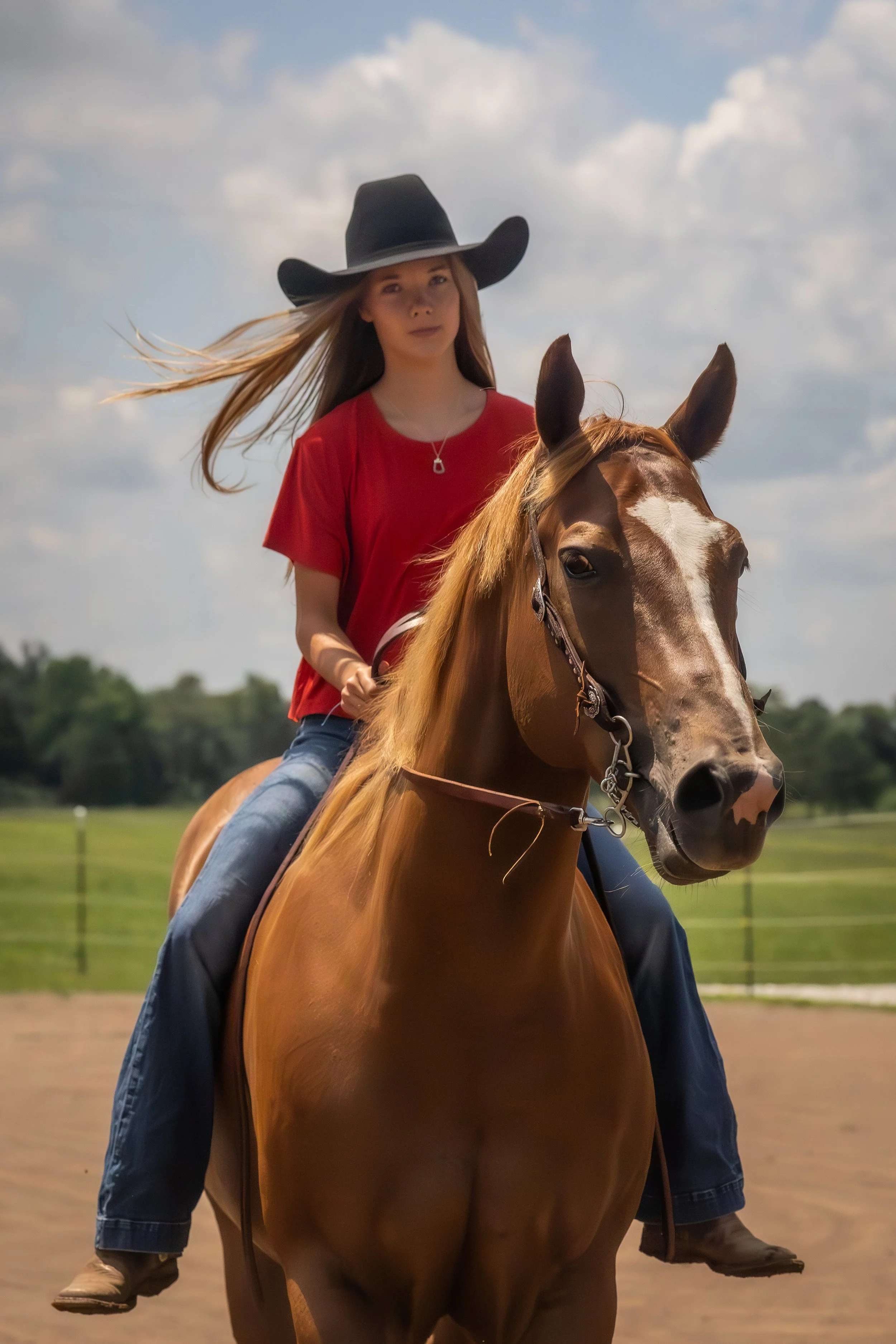 A woman wearing a black cowboy hat and red shirt riding a brown and white horse outdoors on a sunny day with some clouds in the sky.