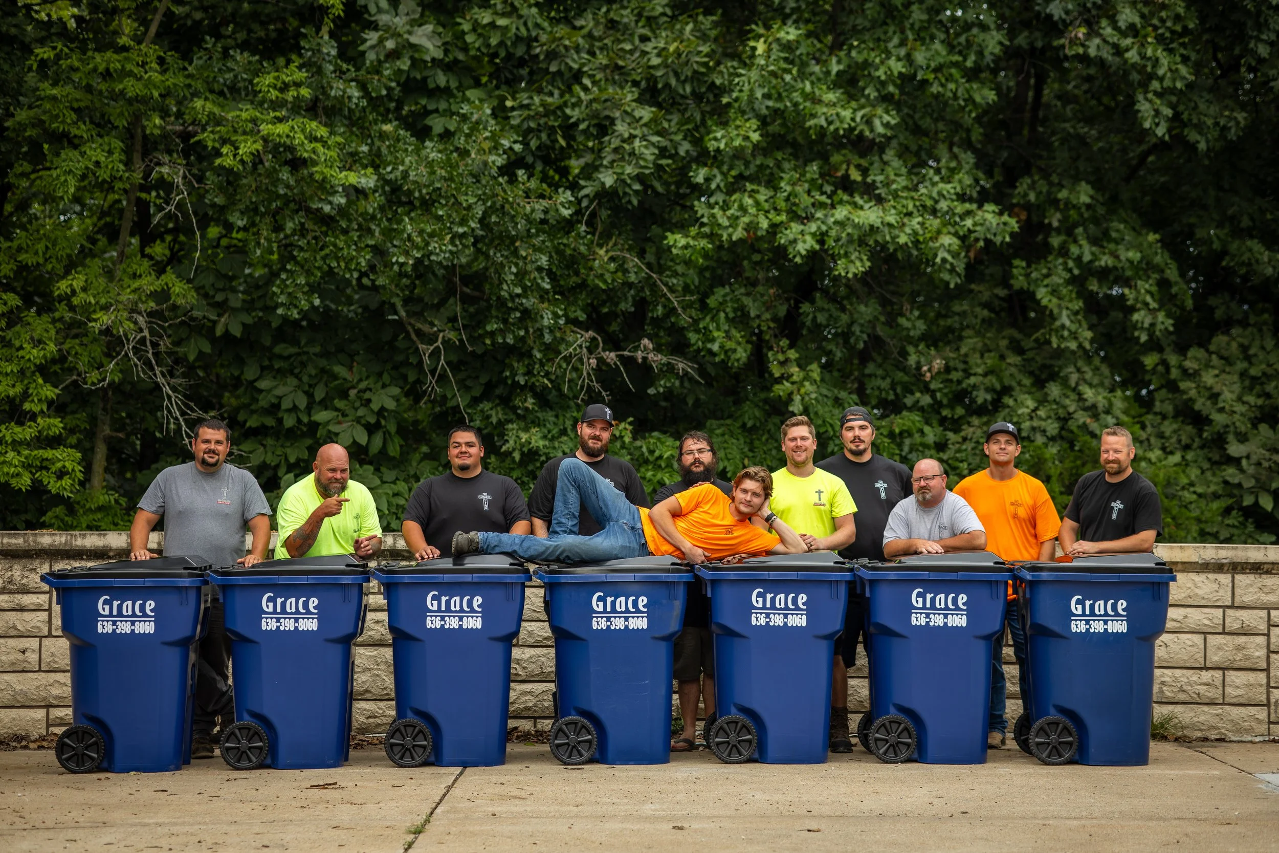 Group of people standing behind blue recycling bins outdoors with dense green trees in the background.