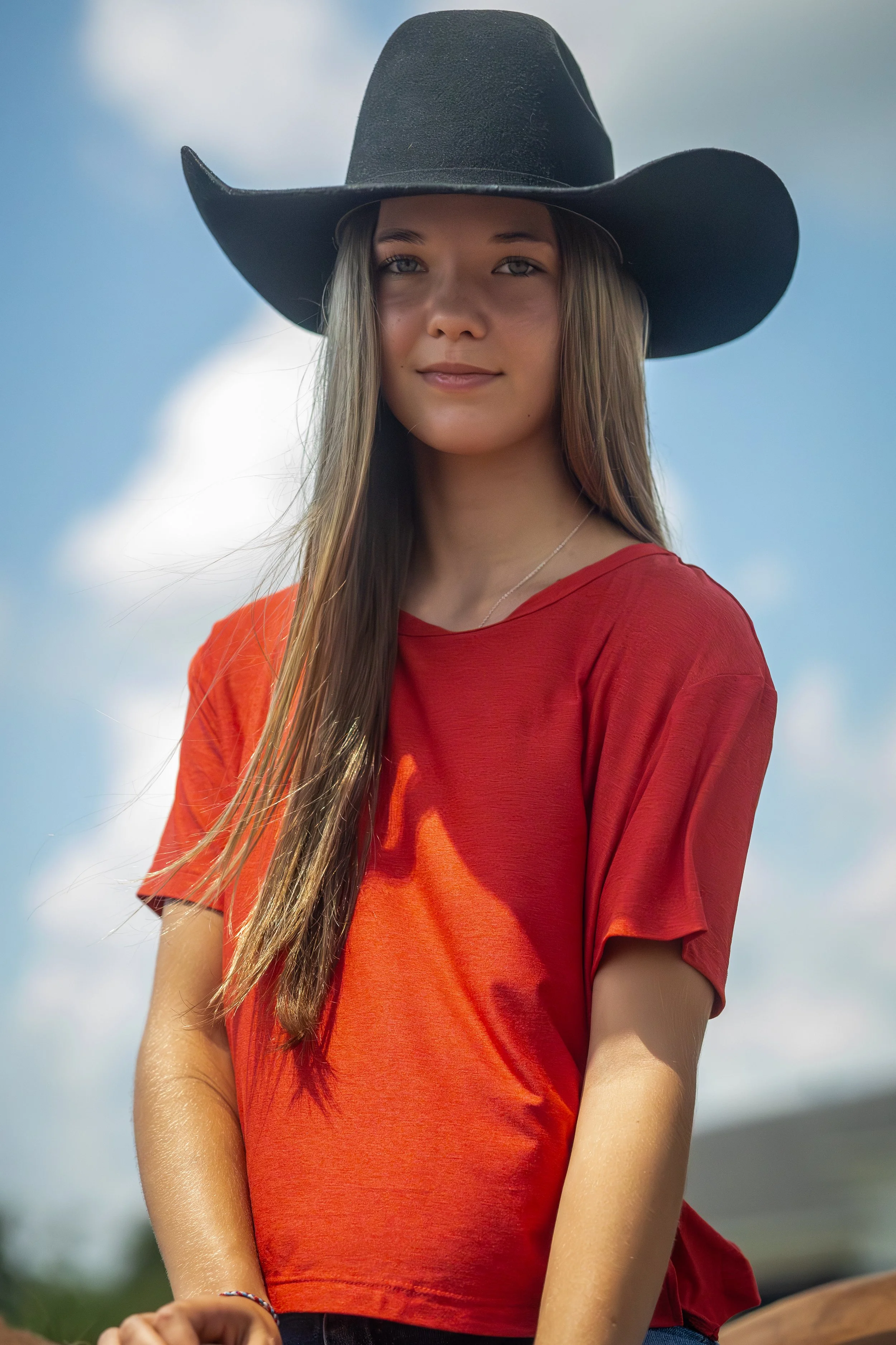 A young woman wearing a large black cowboy hat and a red shirt looks at the camera against a blue sky with scattered white clouds.