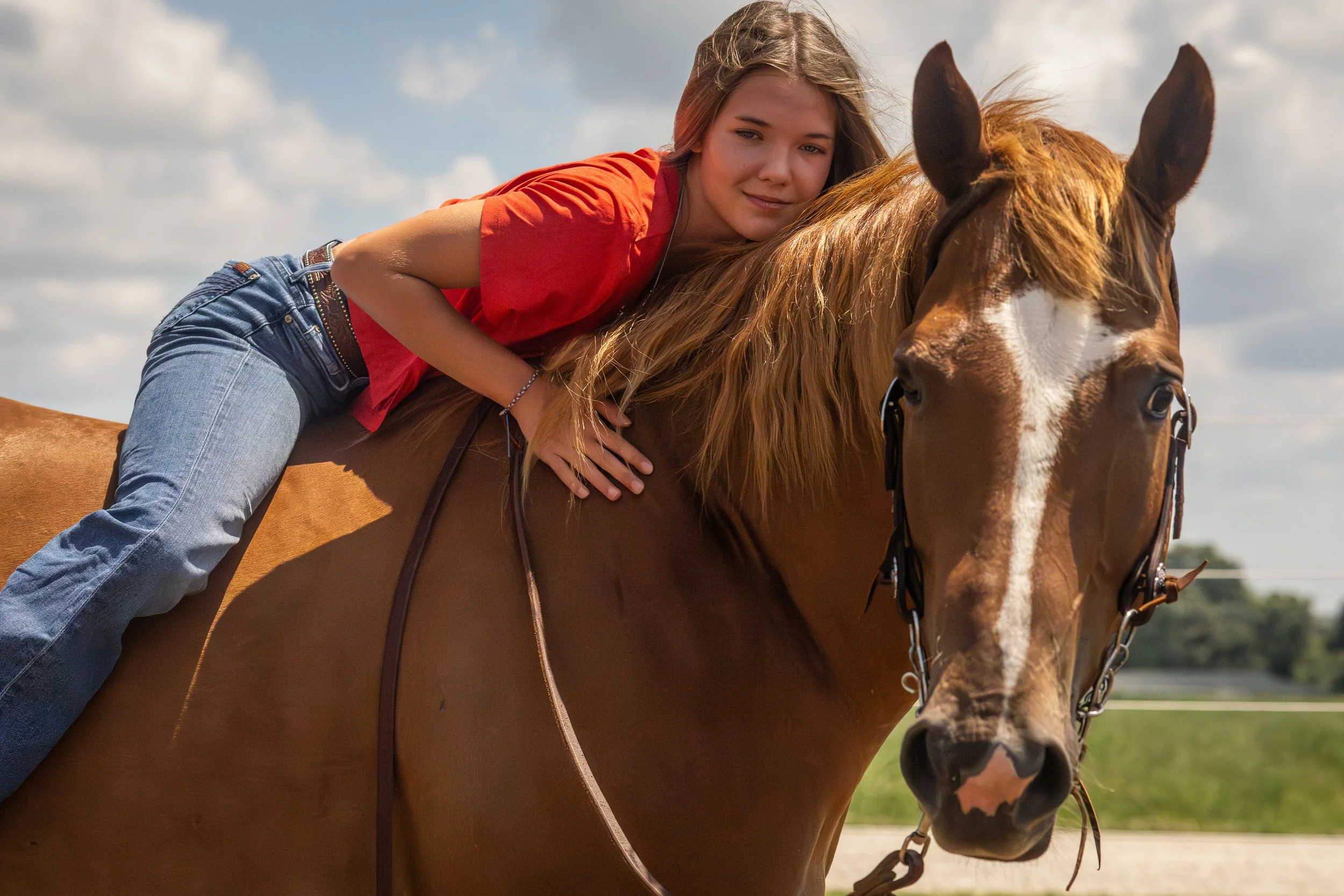 Young woman riding a brown horse outdoors, sky with clouds in the background, wearing a red shirt and blue jeans.