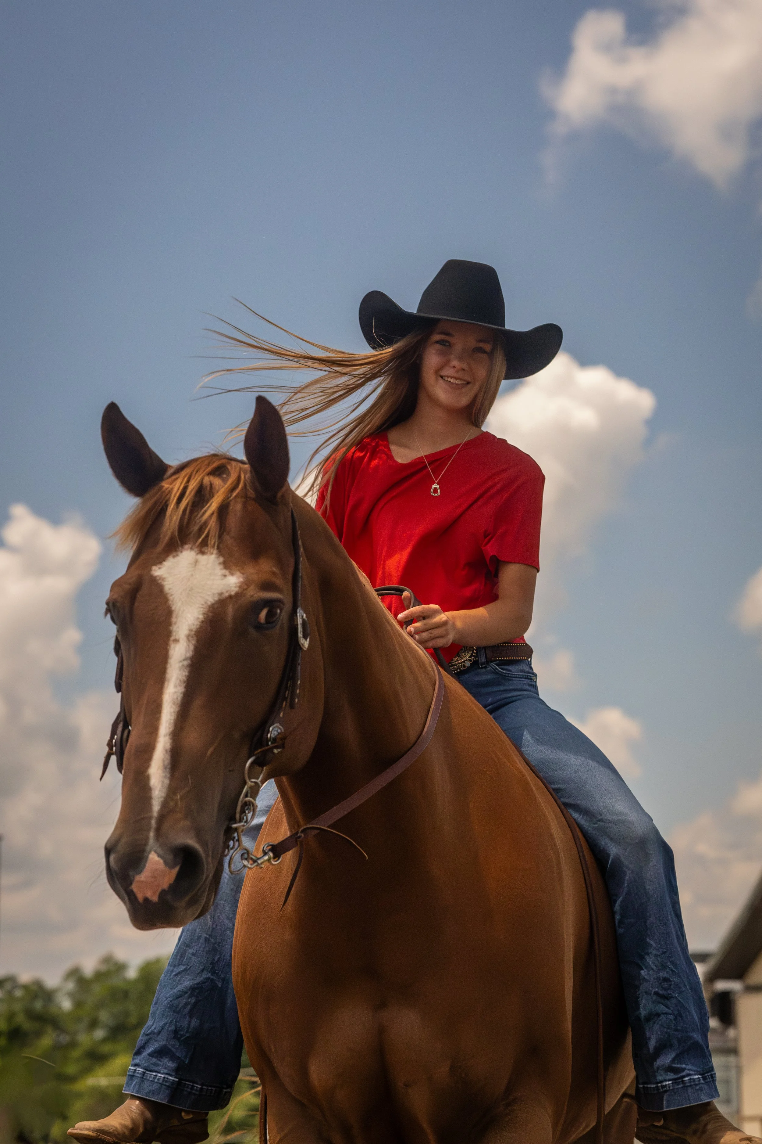 A woman riding a chestnut horse outdoors on a bright day with a blue sky and white clouds, wearing a black cowboy hat and a red shirt.