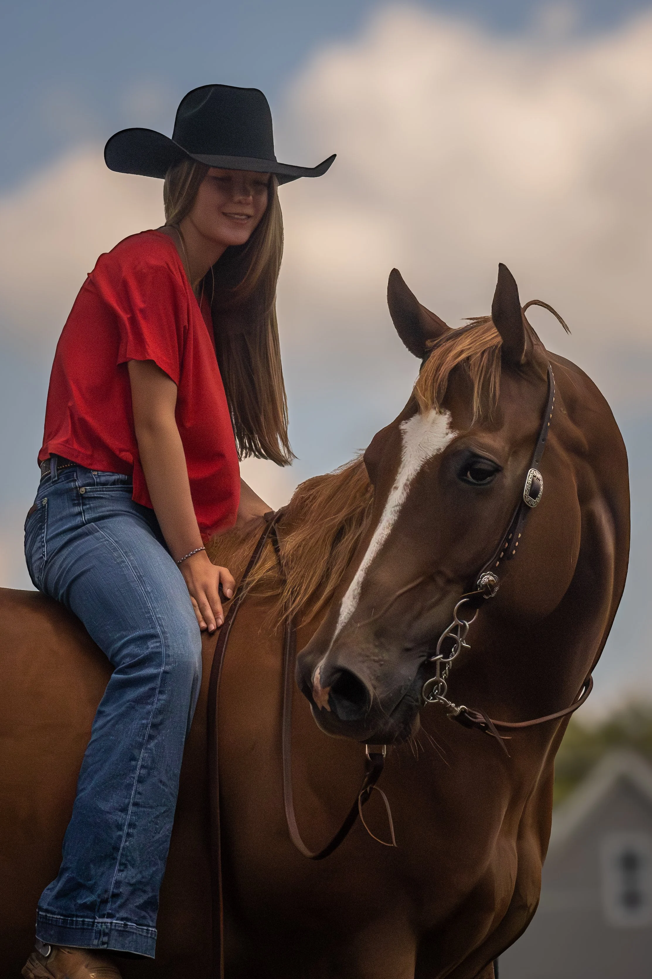 A young woman wearing a red shirt, blue jeans, and a black cowboy hat riding a brown horse with a white stripe on its face, outdoors with a cloudy sky in the background.