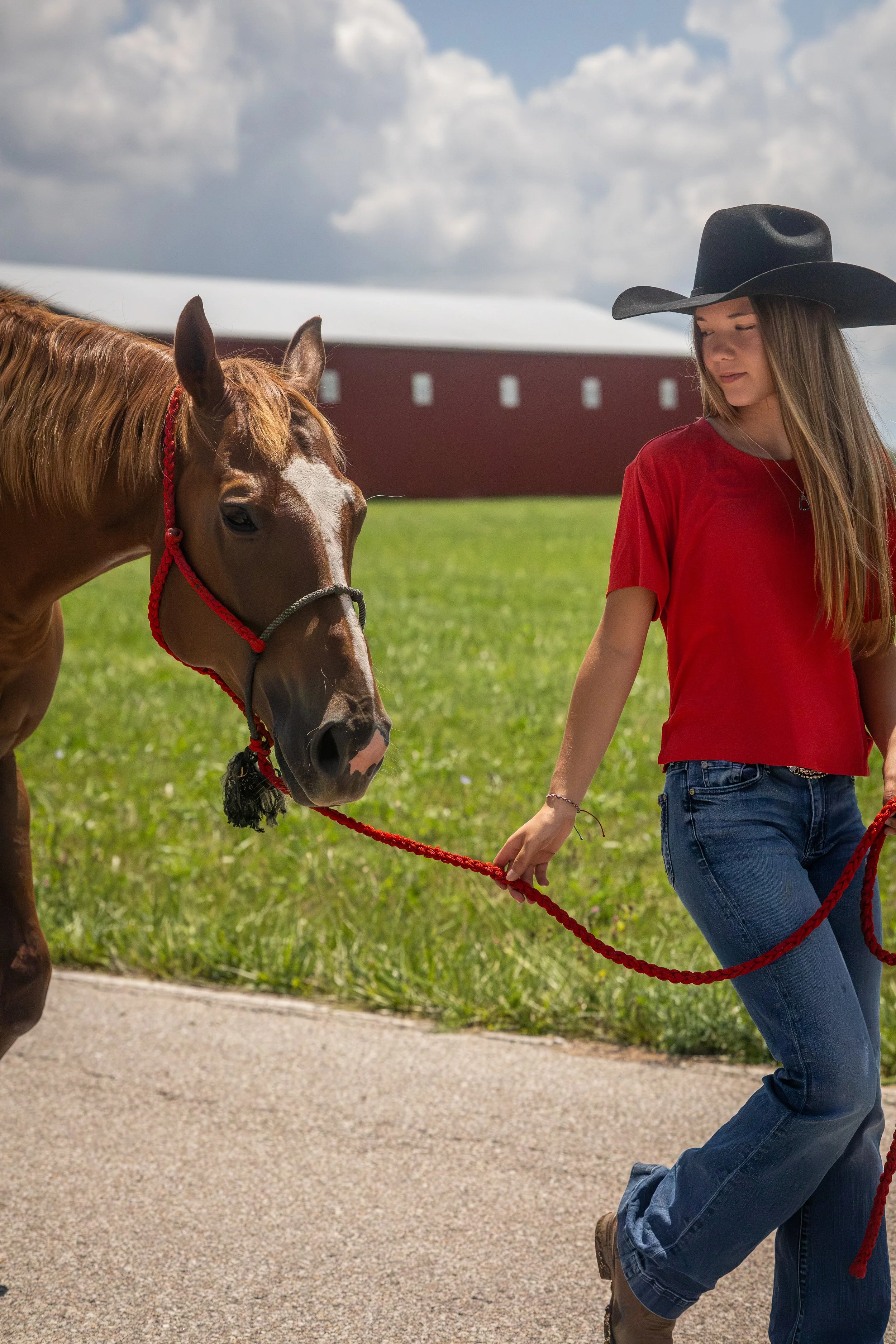 A young woman with long blonde hair wearing a black cowboy hat, red shirt, and blue jeans walking a brown horse with a red halter and a black halter. They are outdoors on a grassy area with a barn and cloudy sky in the background.