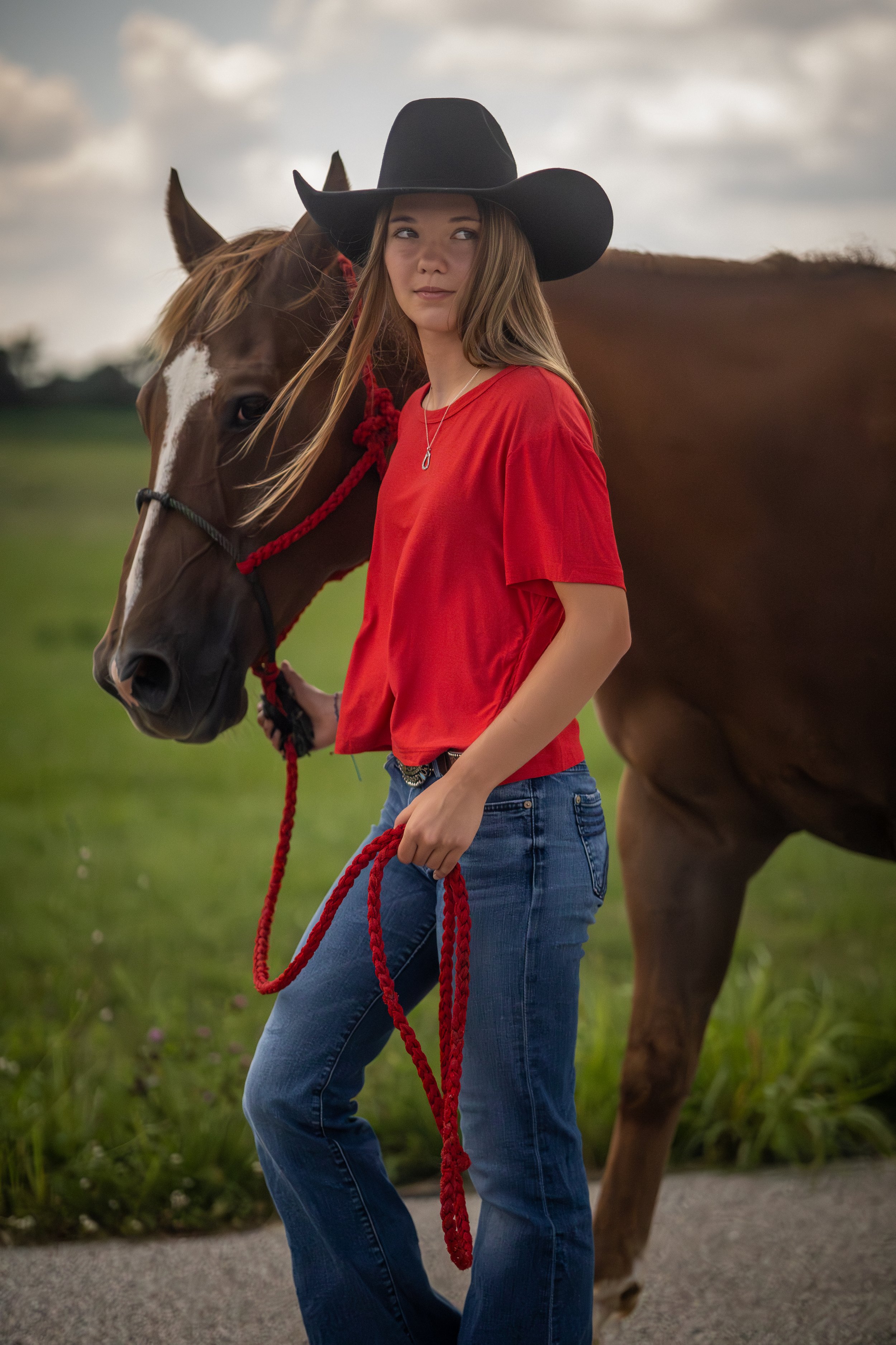 A young woman wearing a black cowboy hat and red shirt standing next to a brown horse with a white blaze on its face in a grassy field during daytime.
