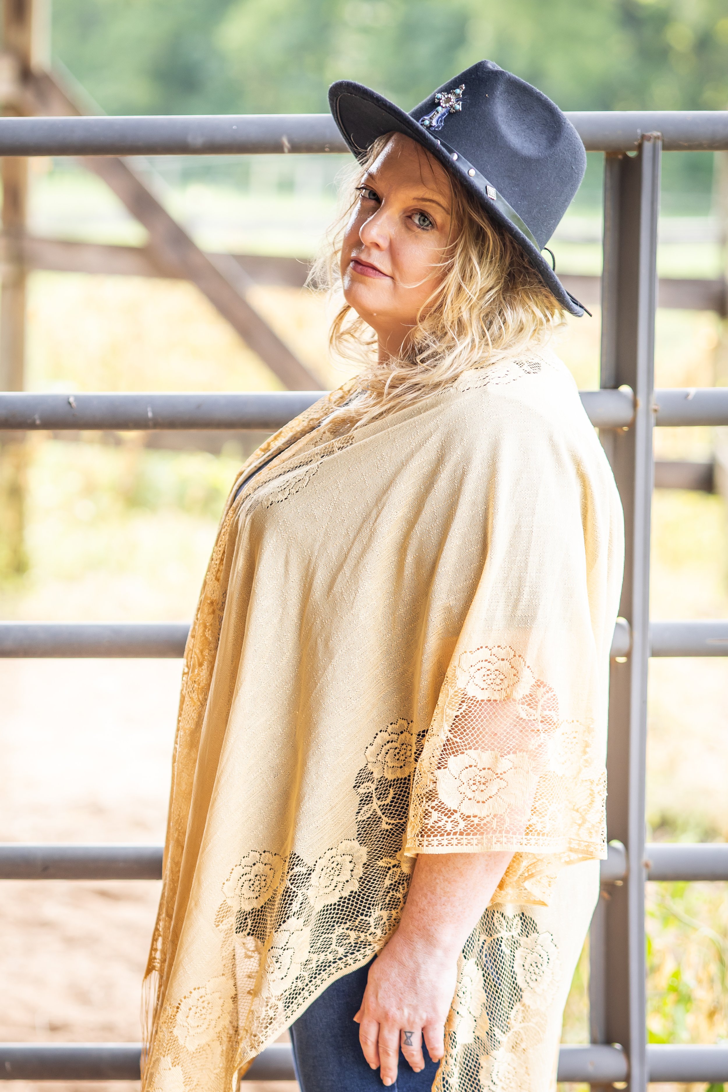 A woman with blonde curly hair wearing a dark gray wide-brimmed hat with decorative pins, a beige lace shawl, and dark pants, standing outdoors near a metal fence during daytime.