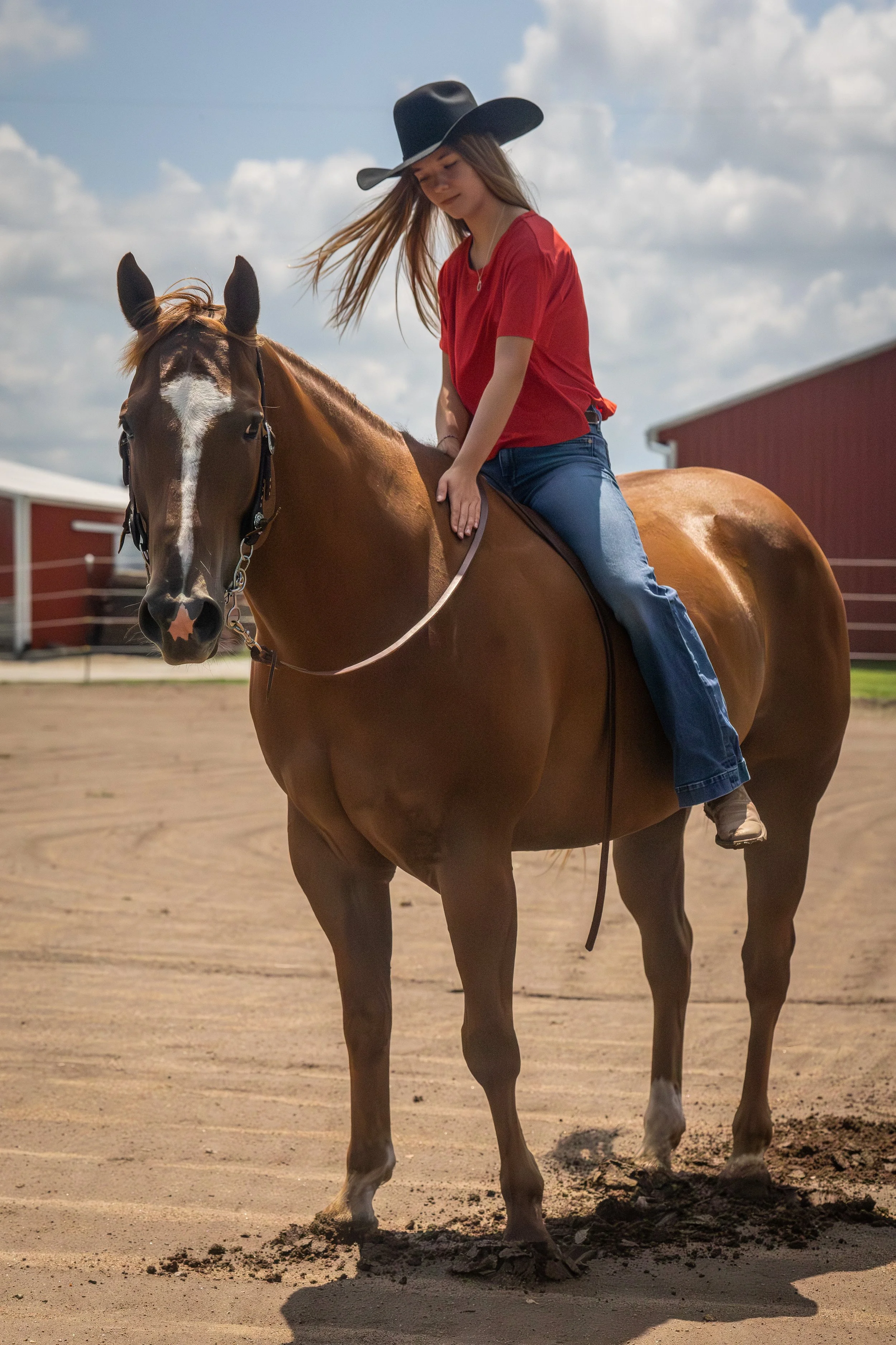 A young woman wearing a black cowboy hat, red shirt, and blue jeans riding a brown horse on a sandy outdoor riding arena with red farm buildings in the background.