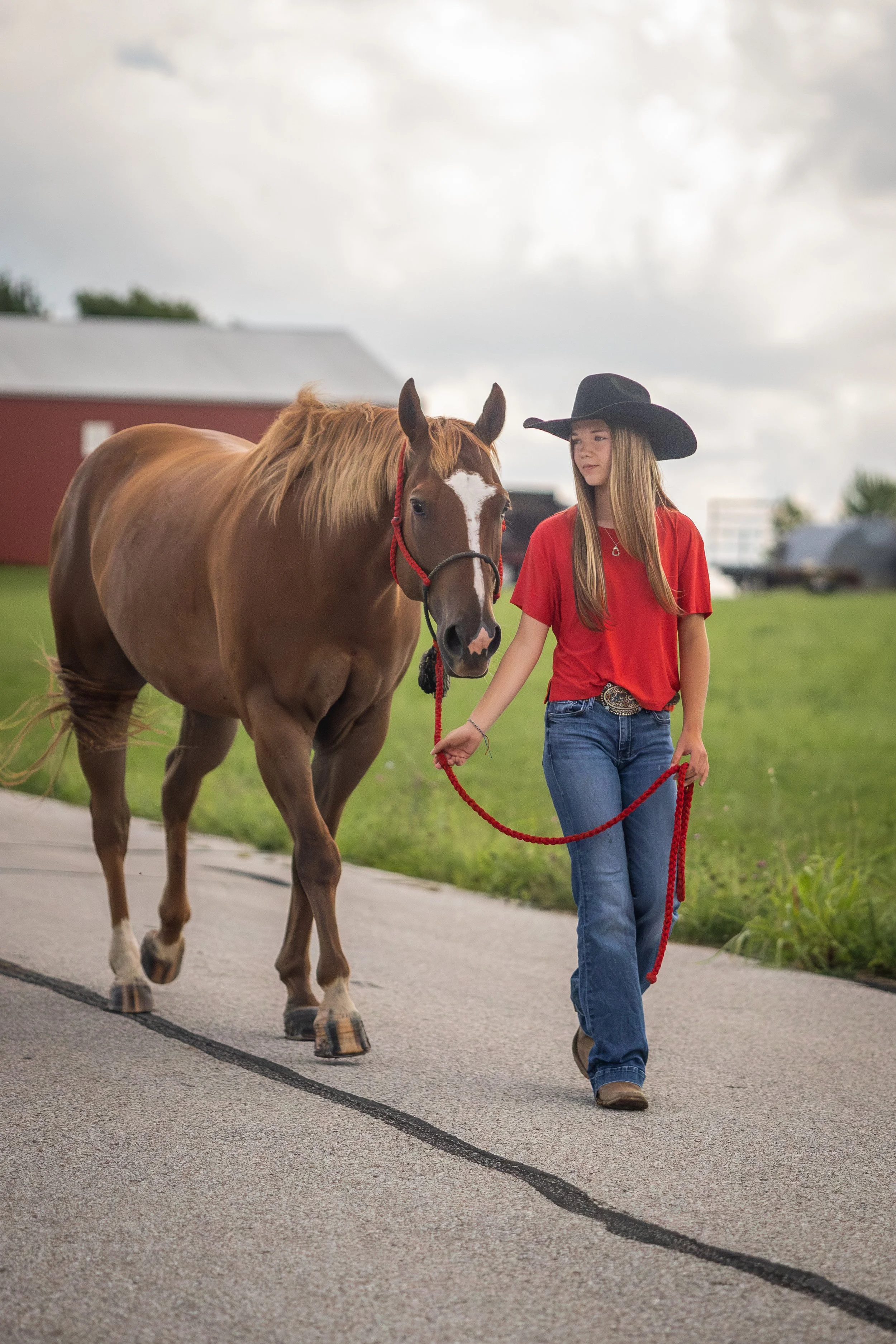 A young woman with long blonde hair, wearing a black cowboy hat, red t-shirt, and blue jeans, walks a brown horse with a white blaze on its face on a paved path in a rural setting. The sky is cloudy, and there is a red barn and green grass in the bac