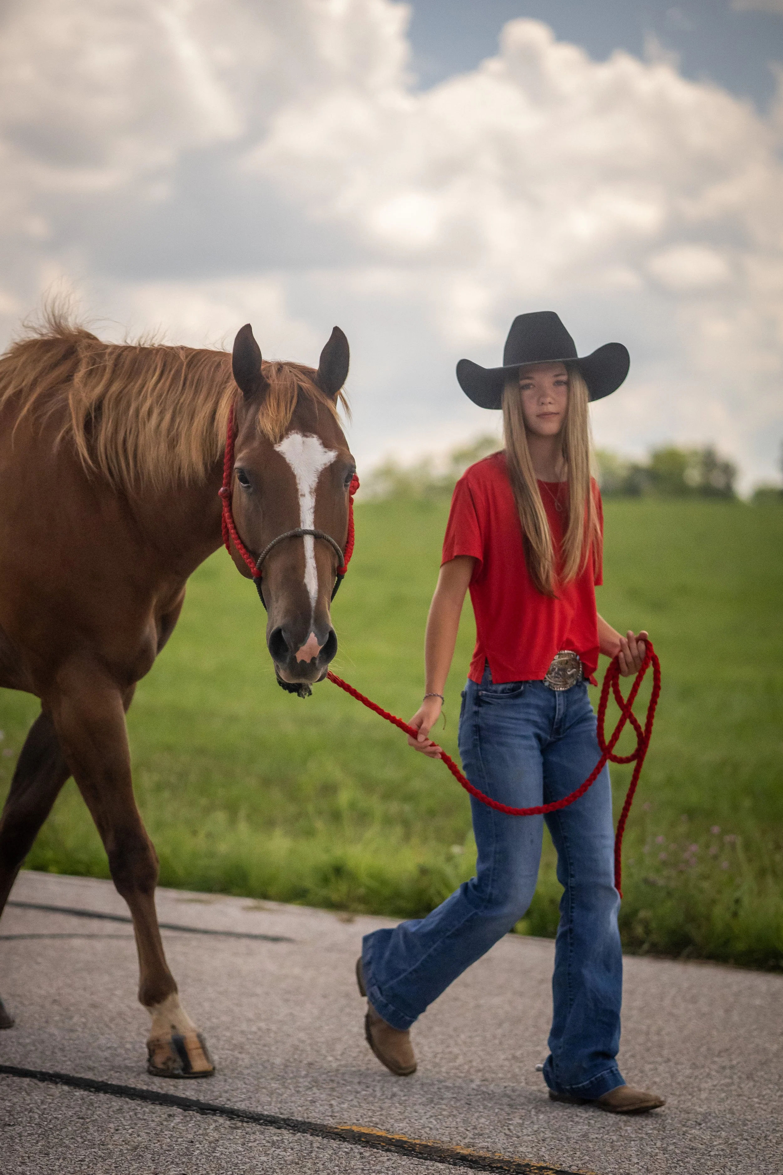 A young woman walking a brown horse with a white star-shaped marking on its face, wearing a black cowboy hat, red T-shirt, blue jeans, and cowboy boots, walking on a paved path in a grassy area under a partly cloudy sky.