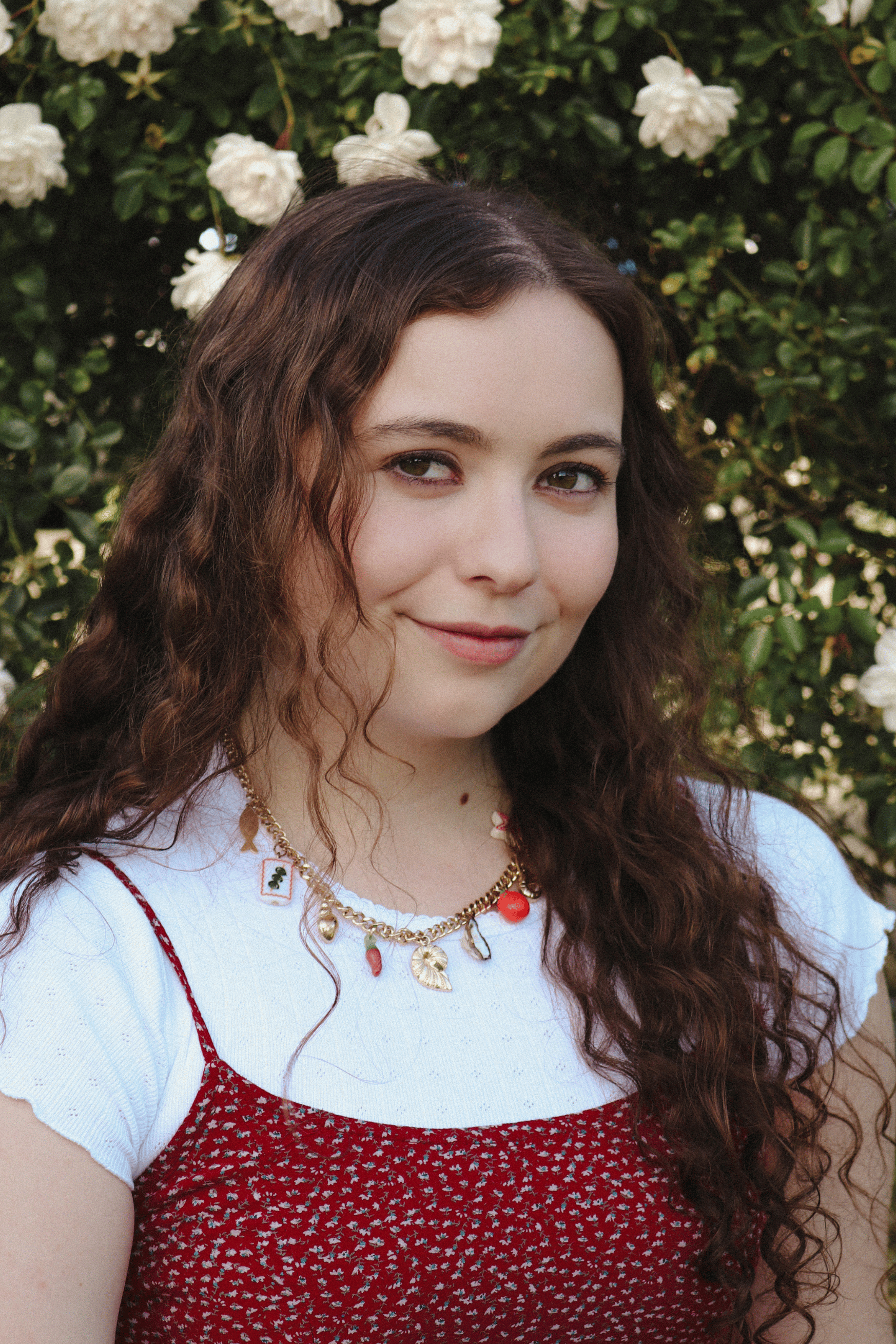 Author Julia Rosemary Turk in a red floral dress with a white undershirt and a charm necklace, with white flowers and green leaves in the background
