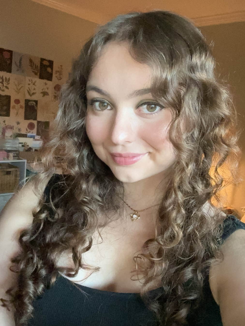 A selfie style headshot of sci-fi fantasy author Julia Rosemary Turk, with curly brown hair and a black top, smiling in a room with flower illustrations on the walls and low warm lighting in the background.