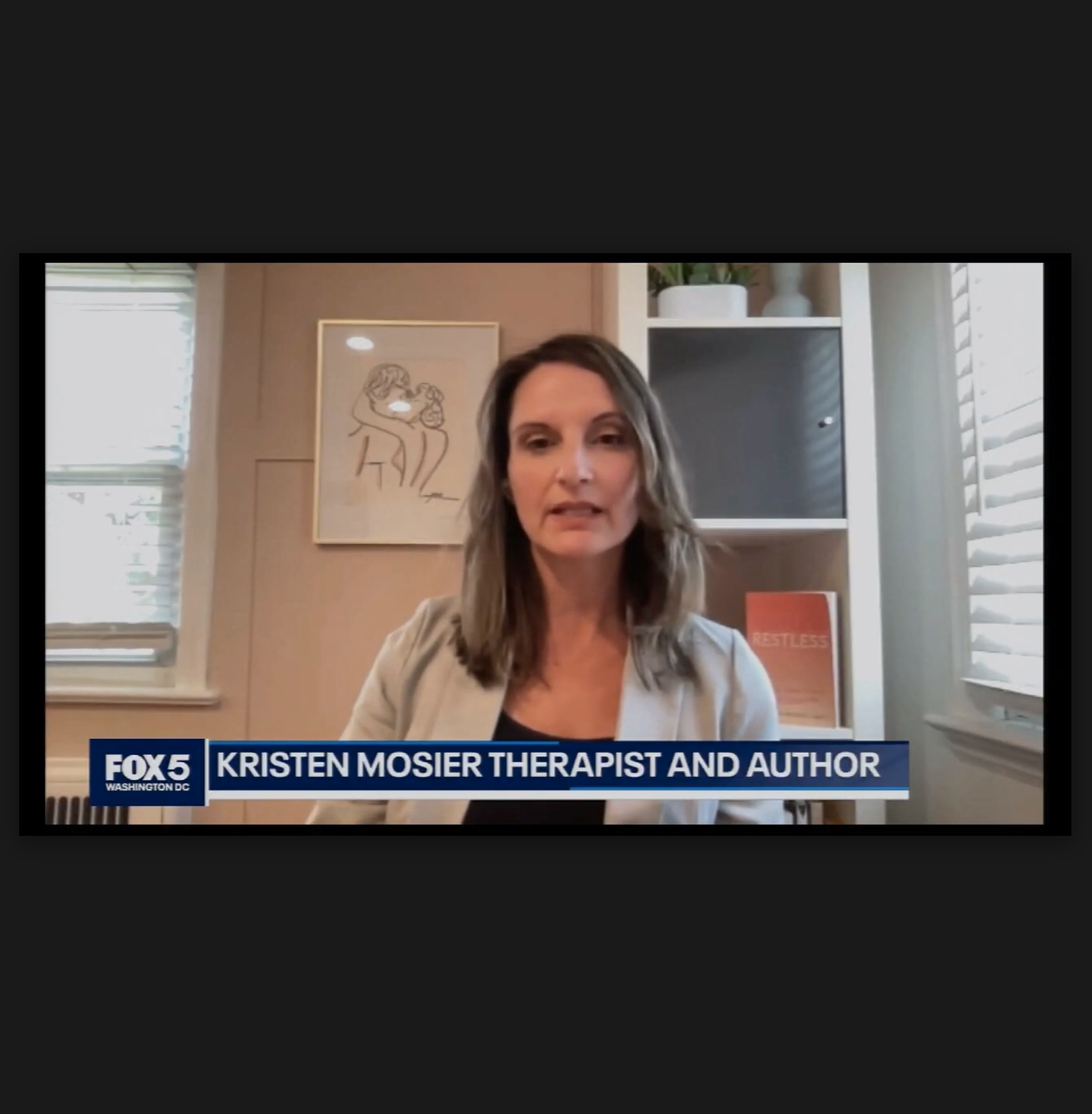 Kristen Mosier, a therapist and author, speaking in a TV interview on FOX5 Washington DC, sitting in a room with a framed artwork of an abstract line drawing of two people. There are windows with blinds on either side and a white shelving unit behind her with books and decorative items.