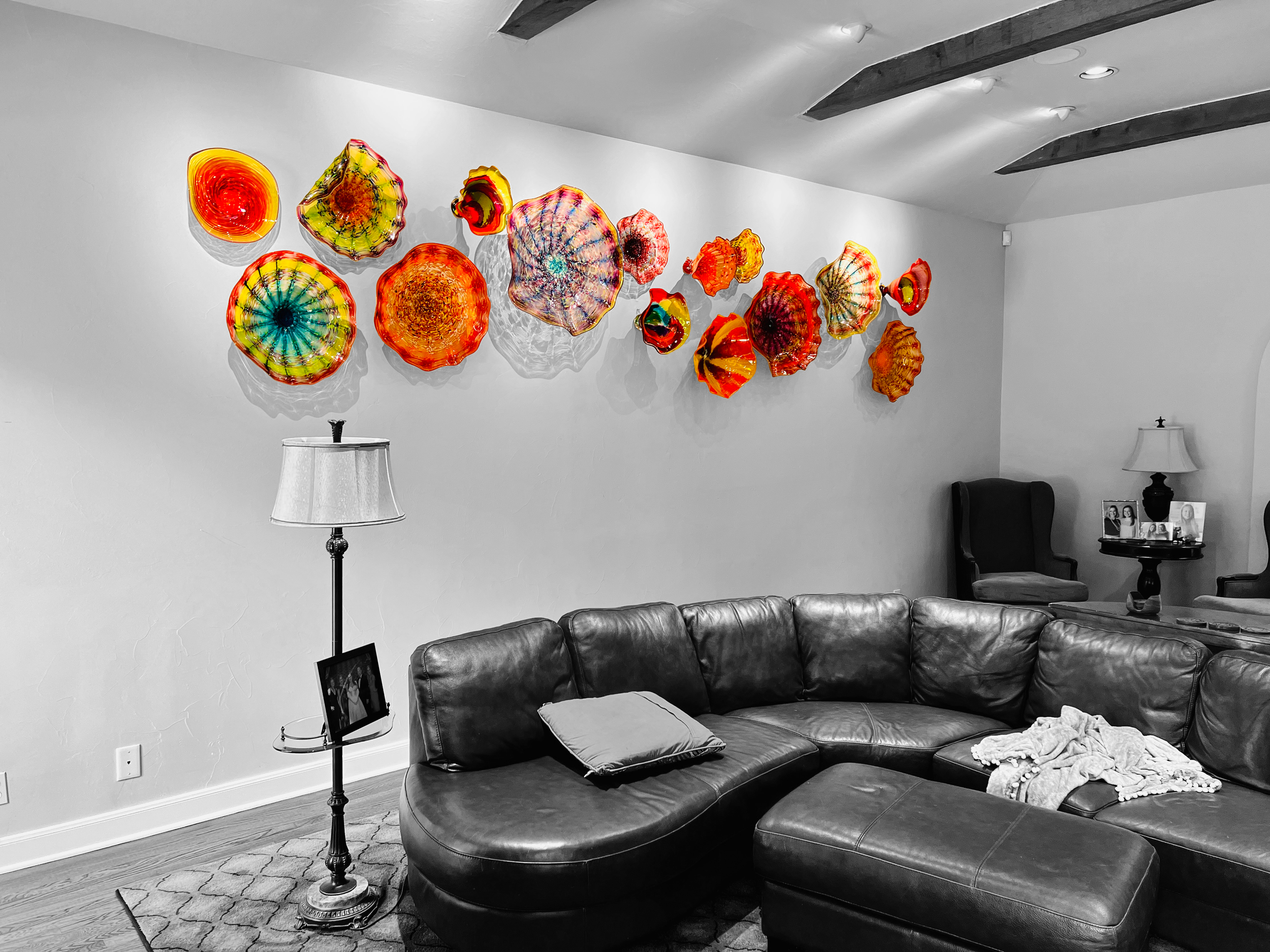 Living room with a black leather sectional sofa, a pillow, and a blanket. There are two table lamps with framed photos on side tables. The wall behind the sofa displays colorful glass art sculptures shaped like sea shells, arranged in a line. The room has wooden floors, a textured rug, and a ceiling with dark wooden beams.