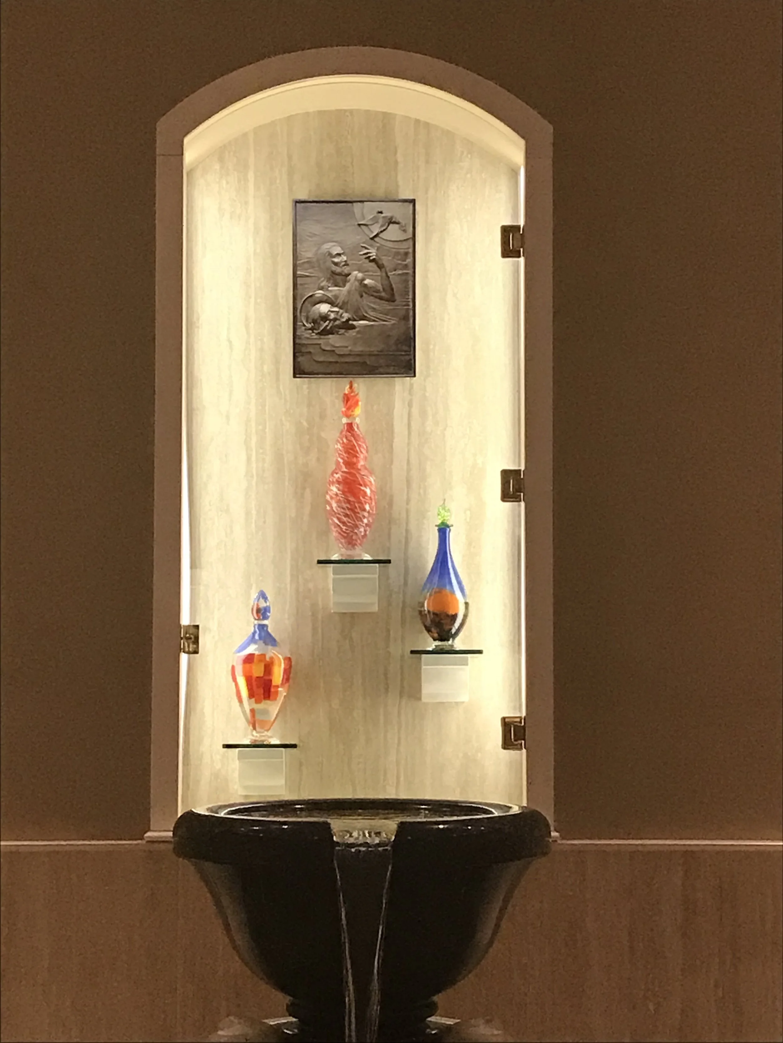 A black fountain in front of a built-in display with three colorful vases and a black and white sculpture of a man holding a fish, inside an arched alcove with lighting.