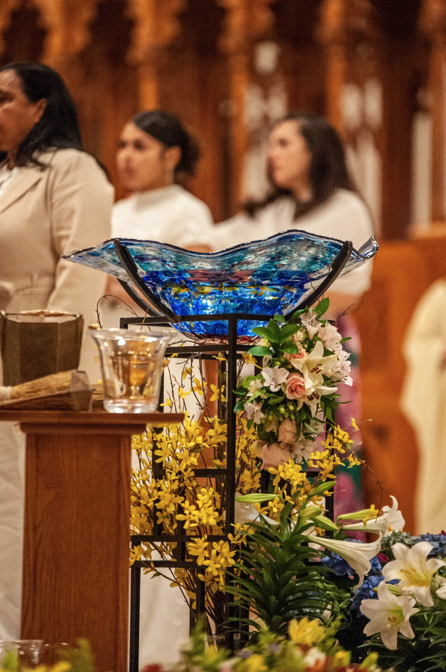 A stained glass bowl with blue, yellow, and red colors on a stand decorated with white, pink, and yellow flowers, with people dressed in white in the background.