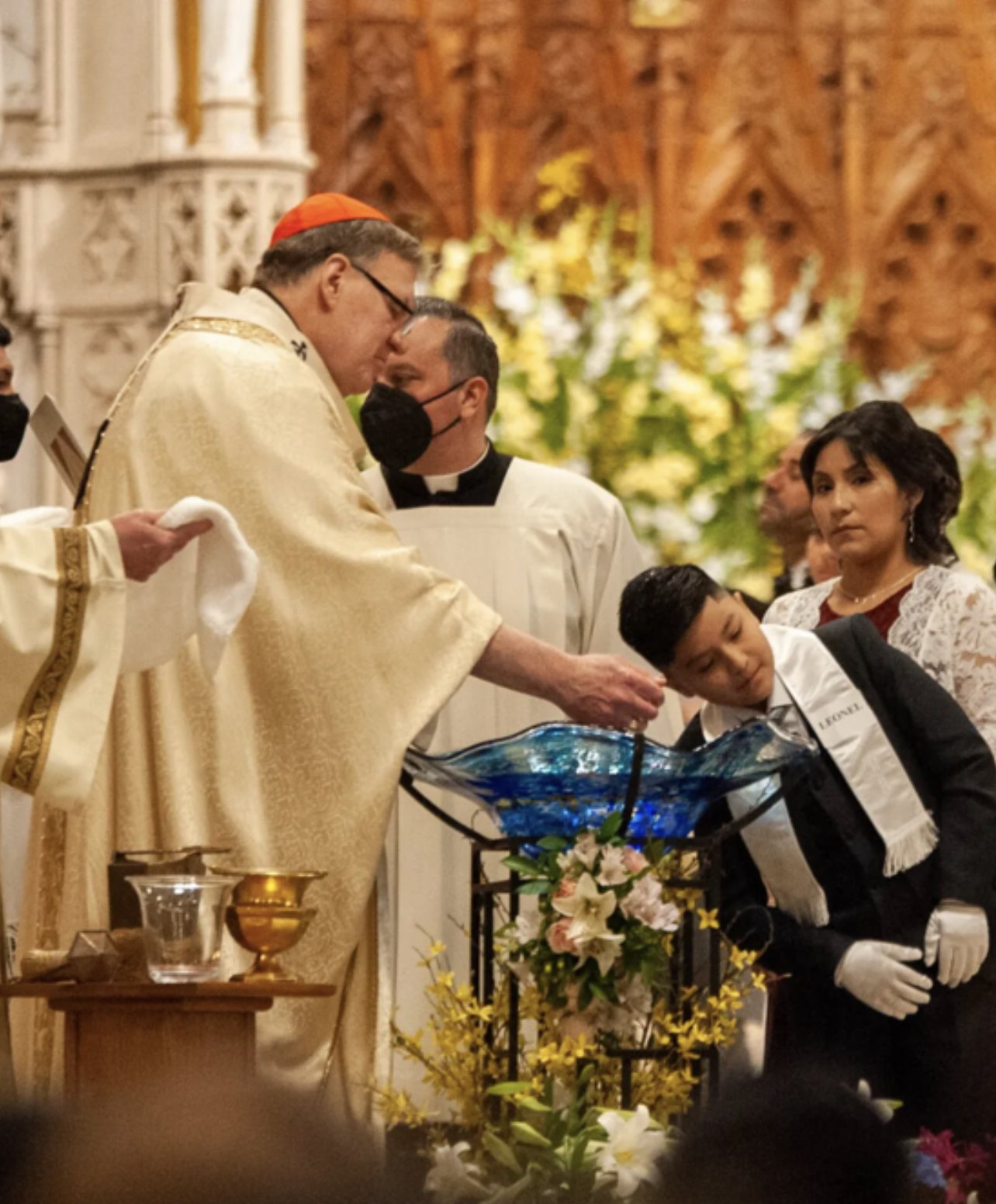 A religious ceremony where a boy is being baptized with water from a decorative glass vessel by a priest dressed in gold robes, with other clergy and adults observing.