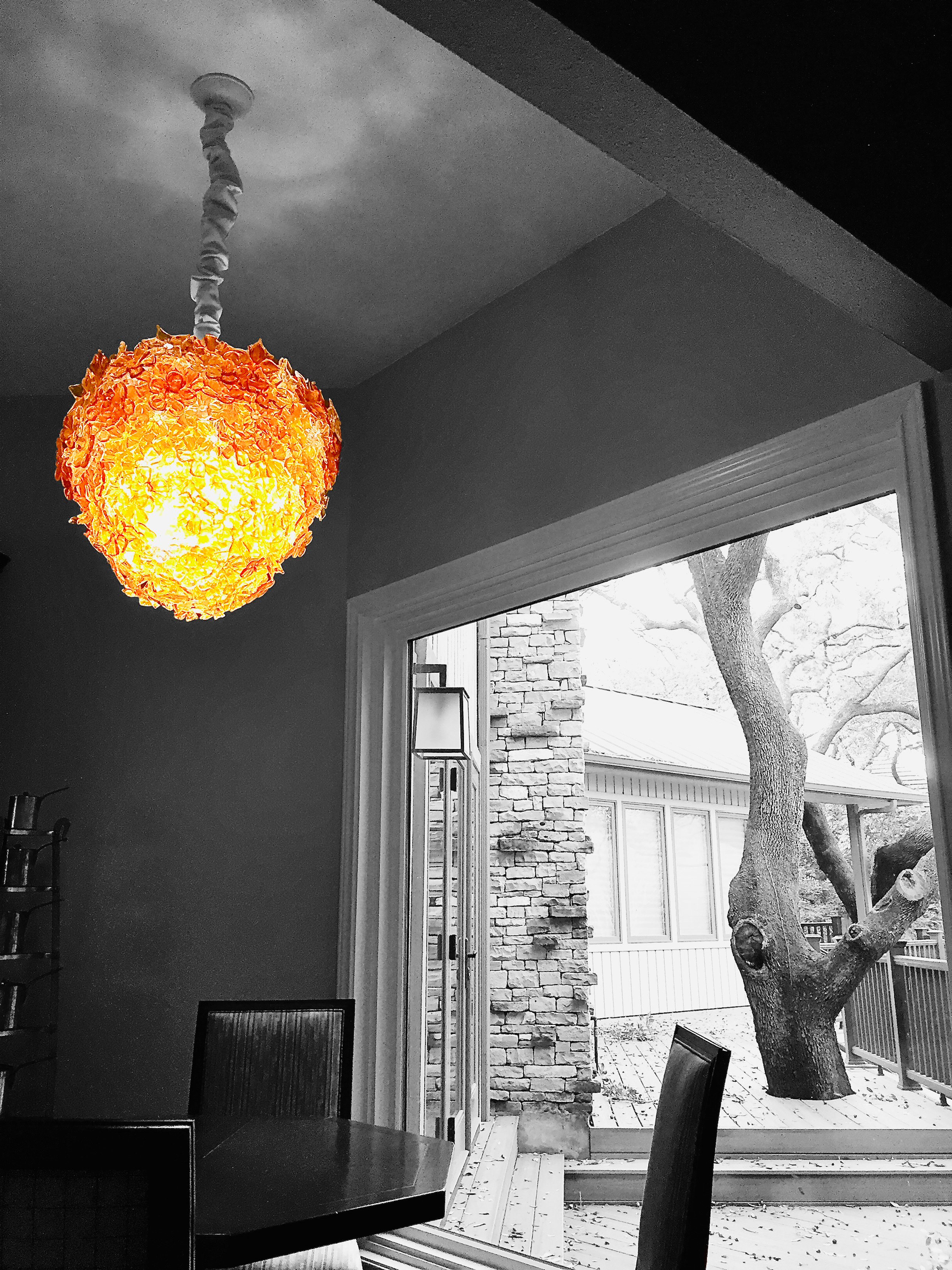A brightly lit, colorful pendant lamp hanging from the ceiling illuminates a dining area, with the rest of the image in black and white, showcasing a wooden dining table and chairs, a sliding glass door, and an outdoor yard with a twisted tree and a brick wall.